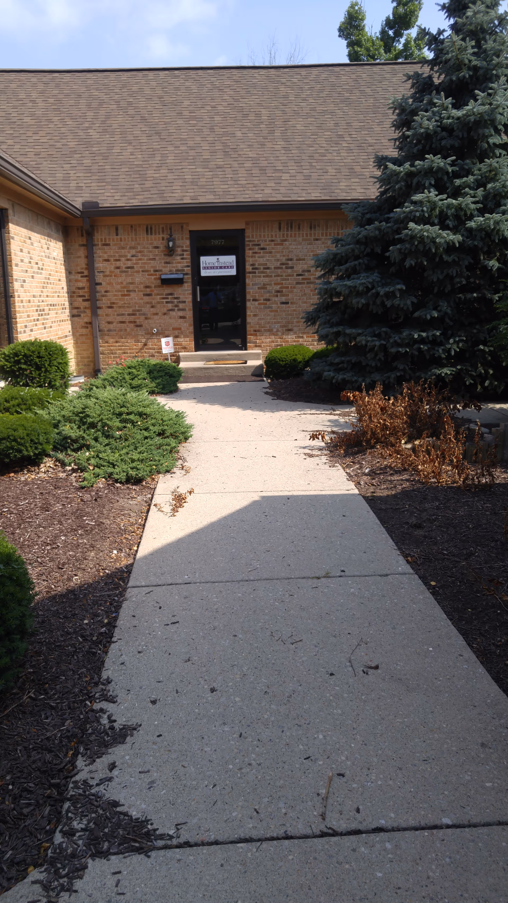Brick exterior of Home Instead facility with entrance path, shrubs, and a tall evergreen tree.