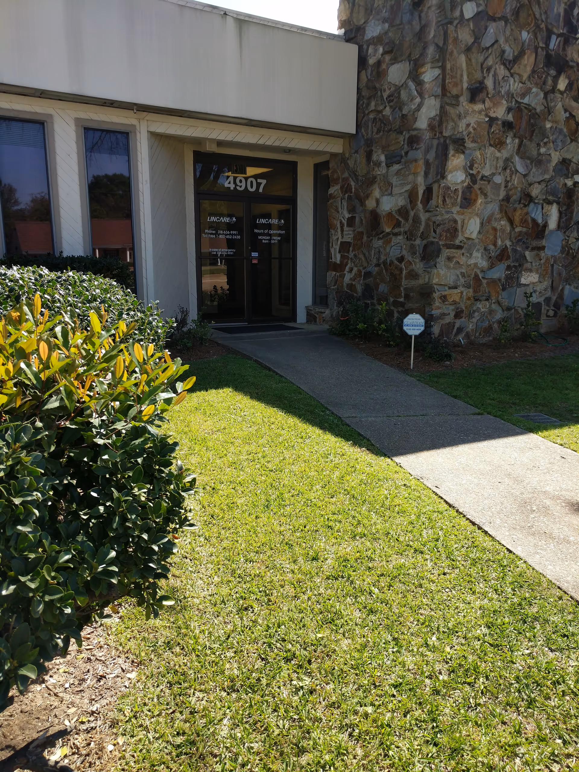Entrance of a LINCARE facility with glass doors, stone wall, shrubs, and a concrete path.