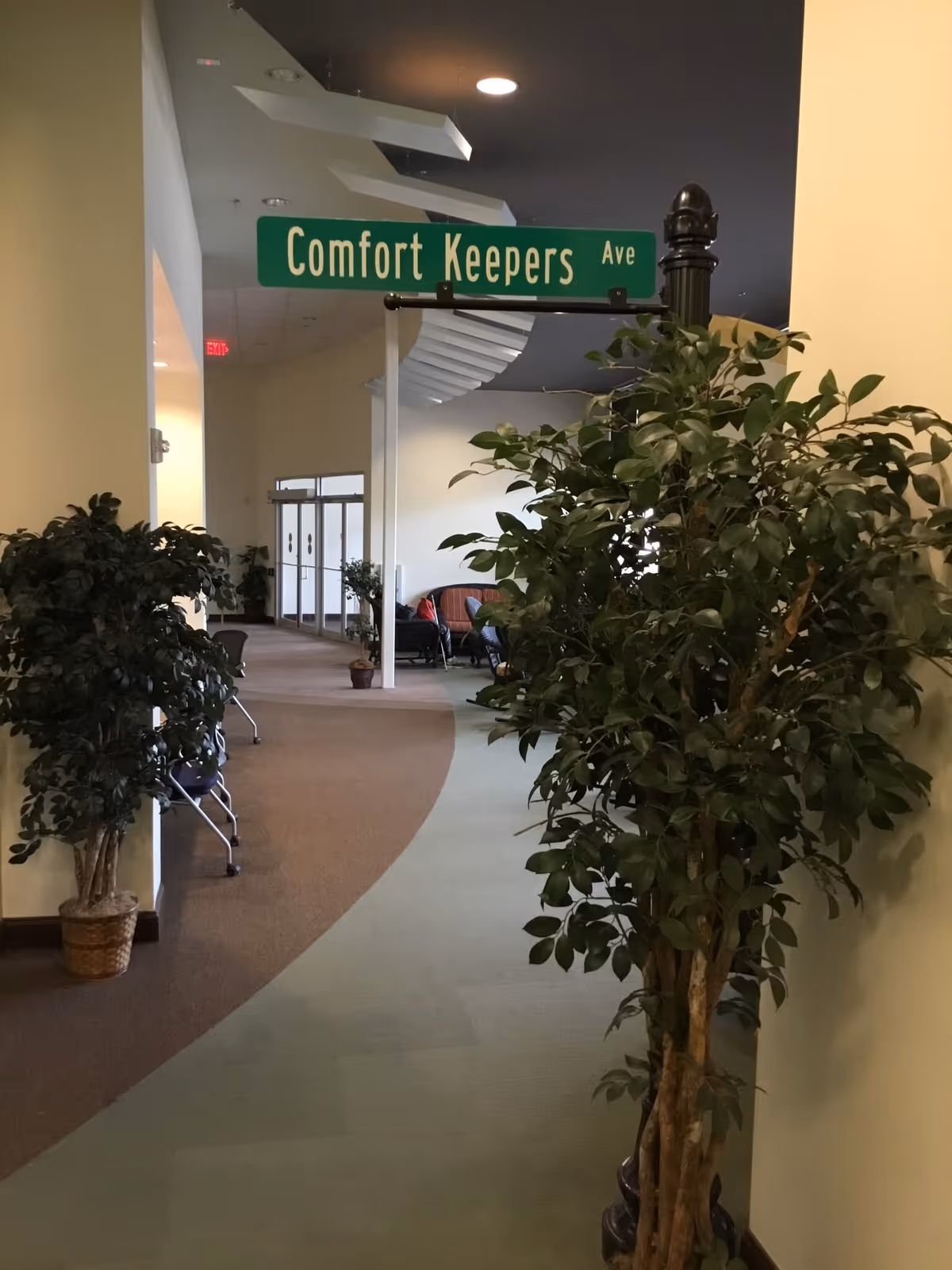 Indoor hallway with green 'Comfort Keepers Ave' sign and potted plants along the corridor.