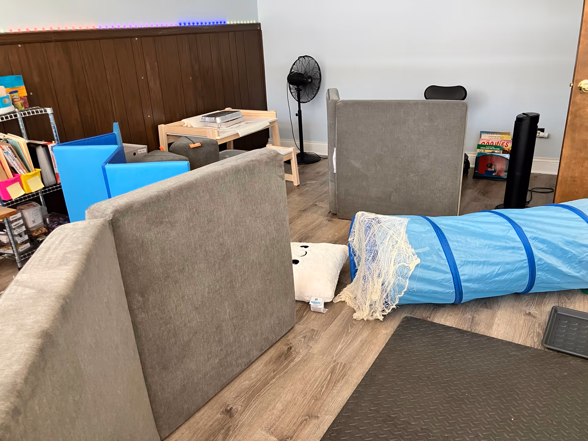 Indoor activity room with gray couches, a blue play tunnel, shelves, and a standing fan.