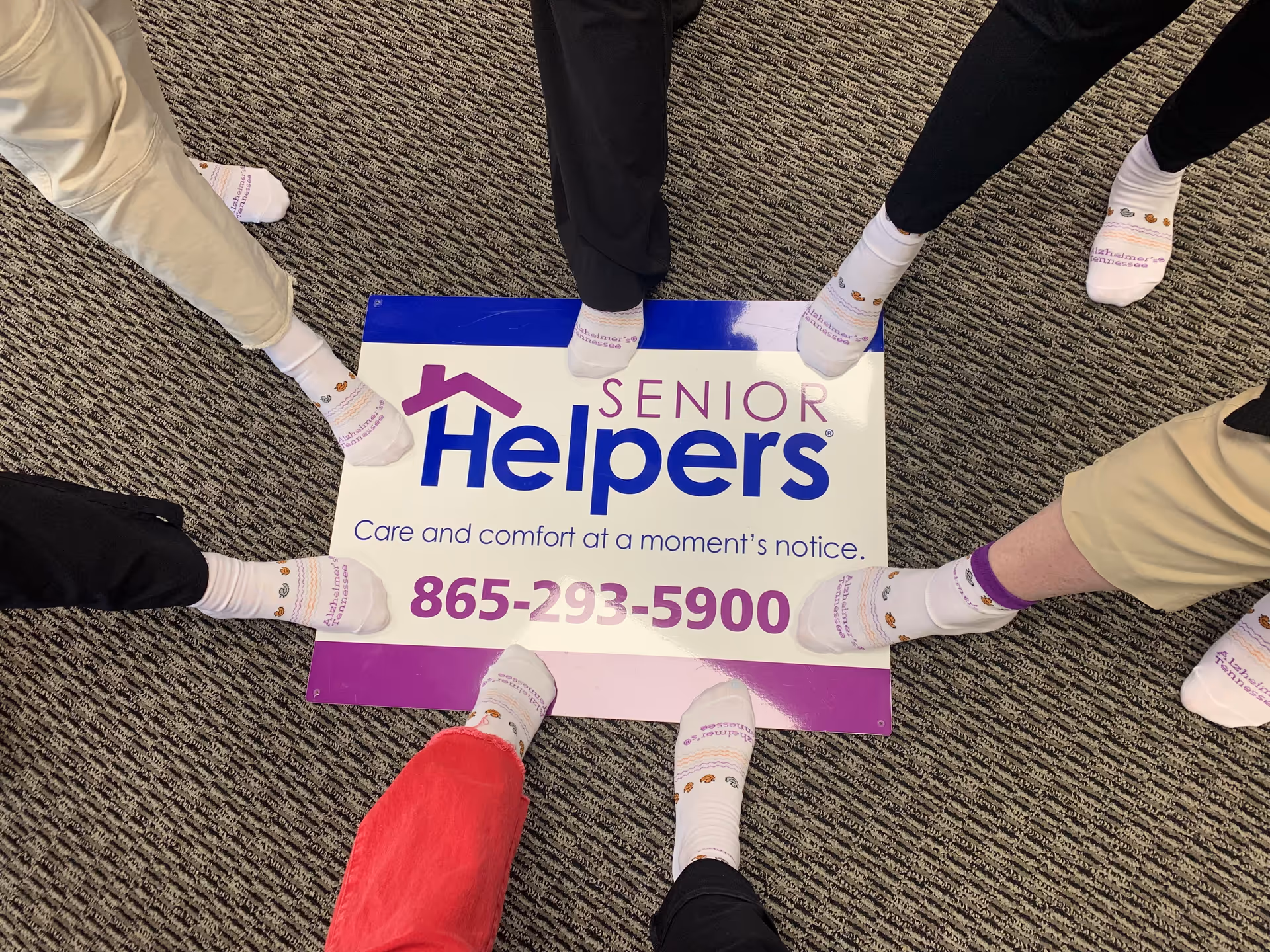 Several people stand around a Senior Helpers sign on a carpeted floor.