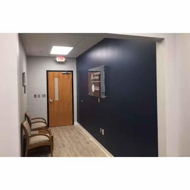 Hallway inside a facility with a dark blue wall, chairs, and a wooden door.