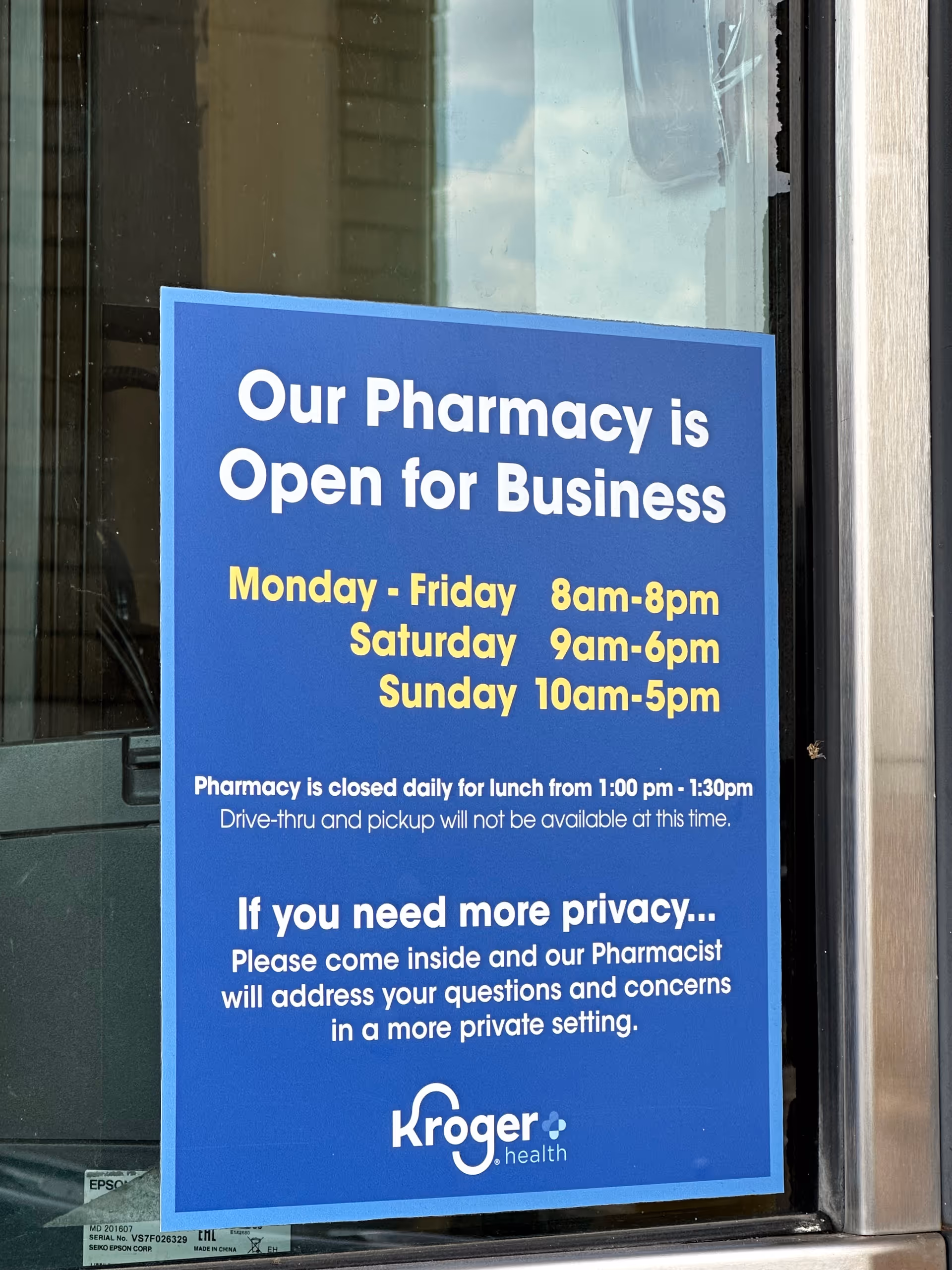 Blue sign in a storefront window reads Our Pharmacy is Open for Business at Kroger Pharmacy.