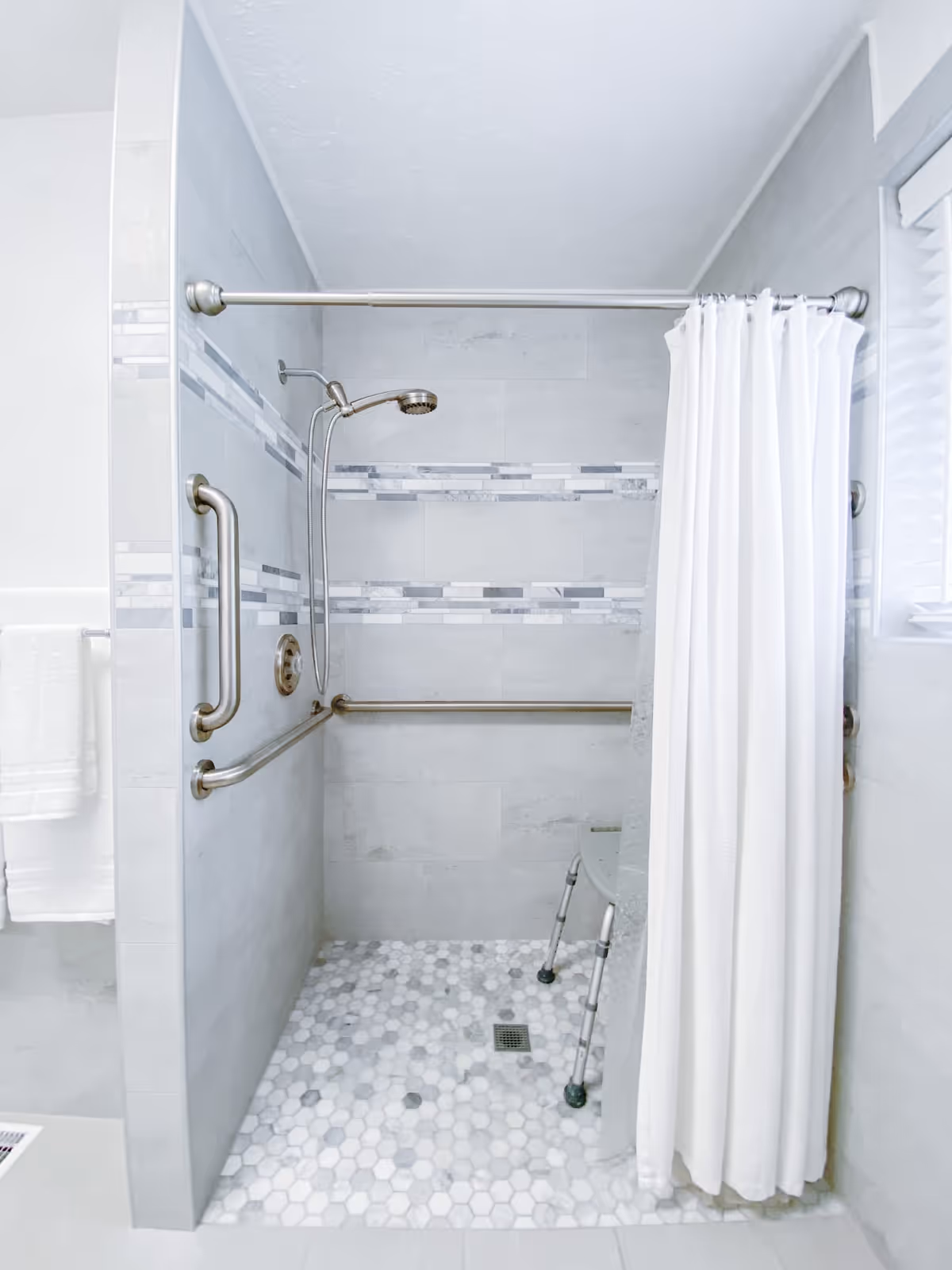 Accessible tiled shower with grab bars, a folding seat, and a white curtain.