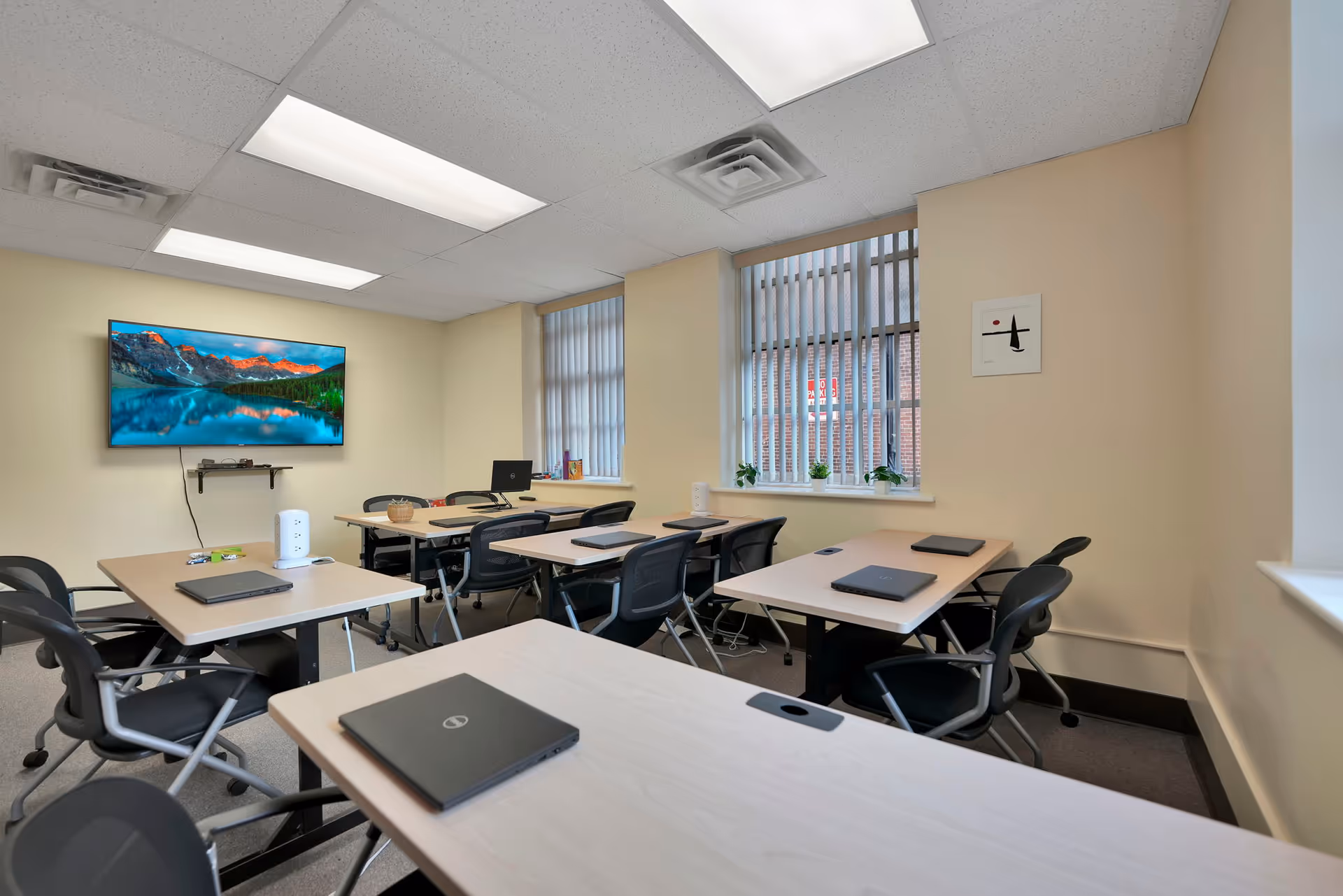 Bright classroom-style meeting room with desks, chairs, laptops, and a wall-mounted TV.