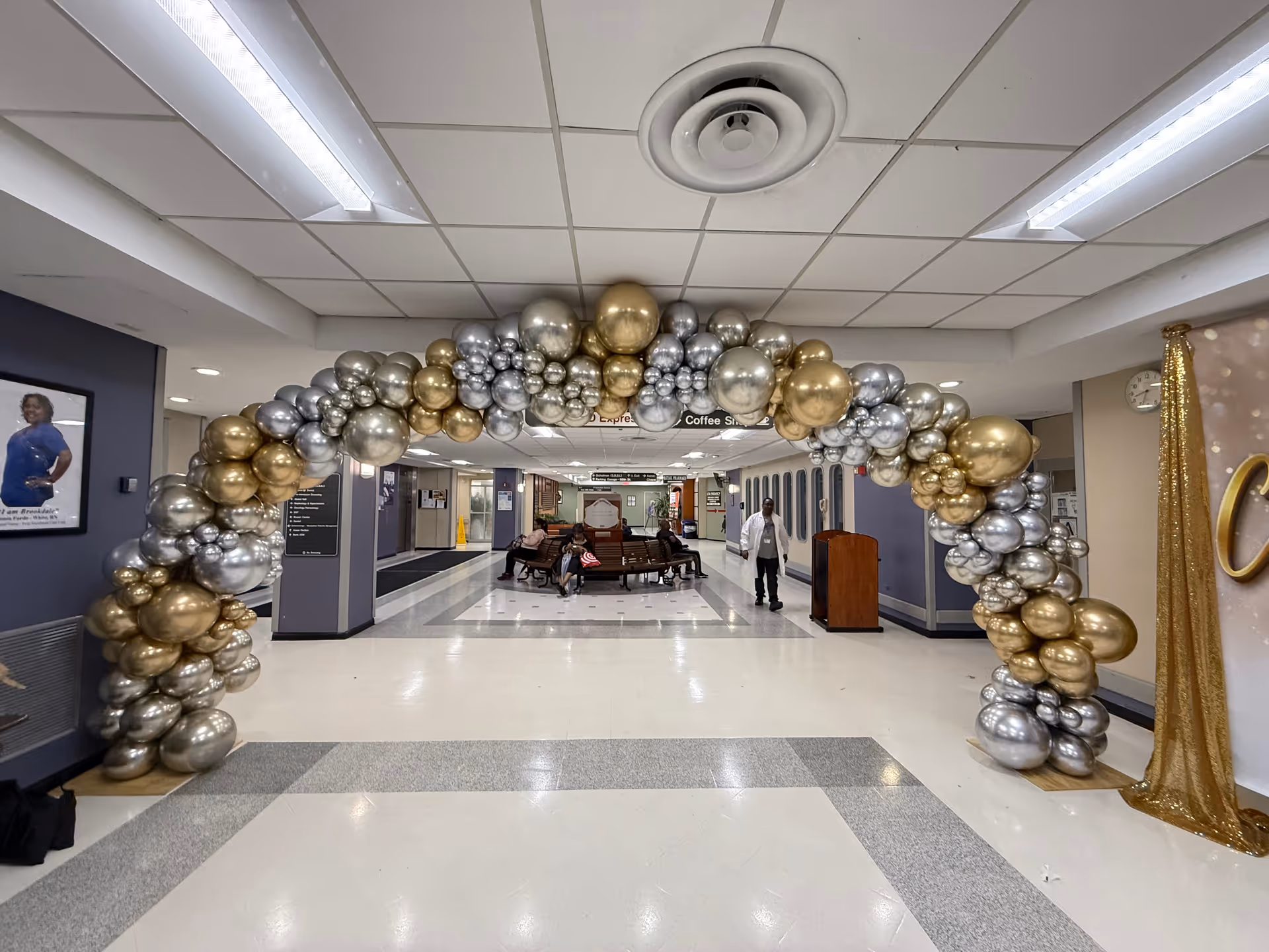 Hospital corridor decorated with a silver-gold balloon arch and seated residents.