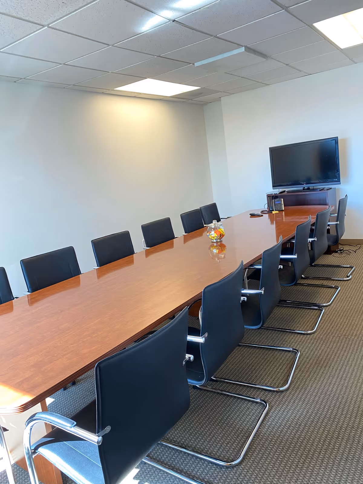 Long conference table with black chairs and a TV in a bright meeting room.