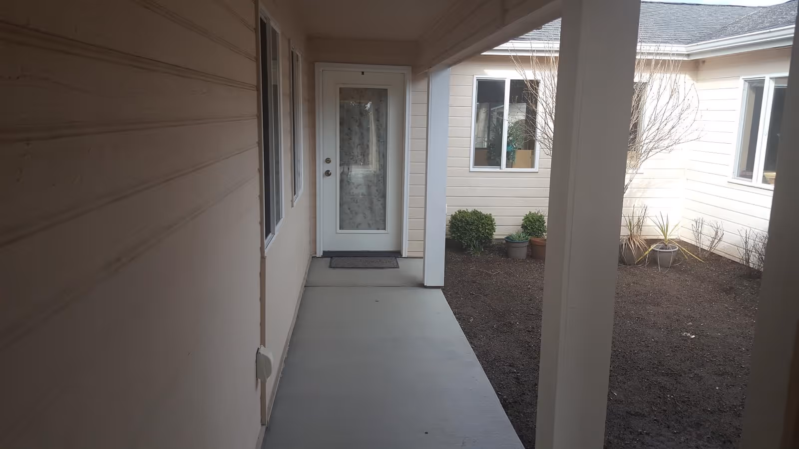 Covered outdoor hallway leading to a door, with windows and potted plants along the path.