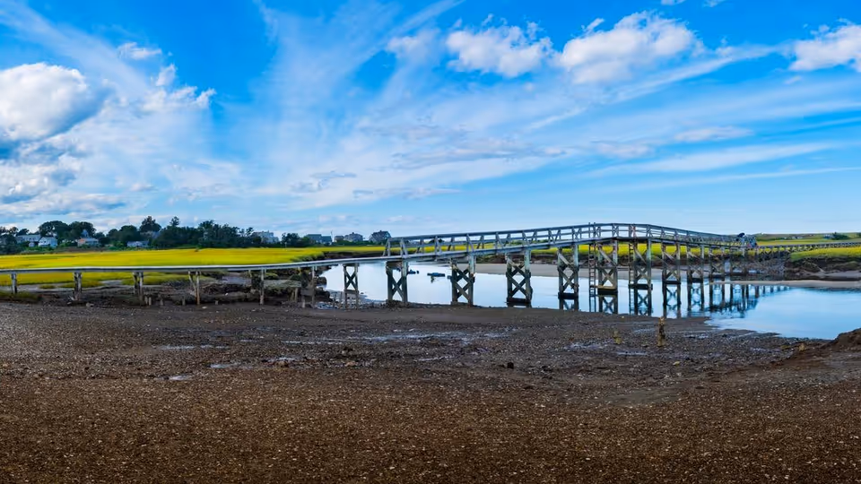 A wooden bridge spans a calm river in a rural landscape under a blue sky.