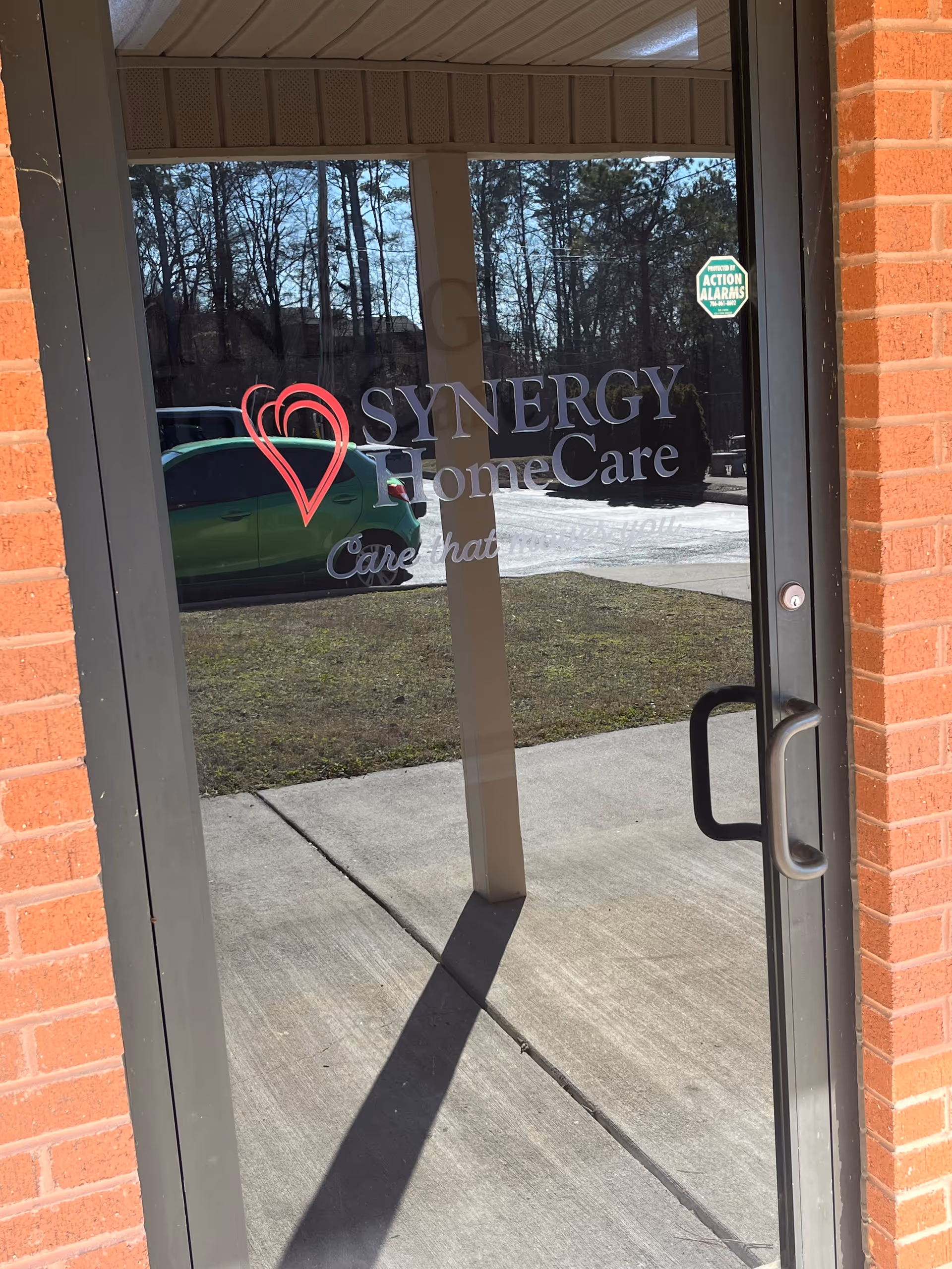 Glass entrance door with SYNERGY HomeCare logo, brick frame, reflection of trees outside.