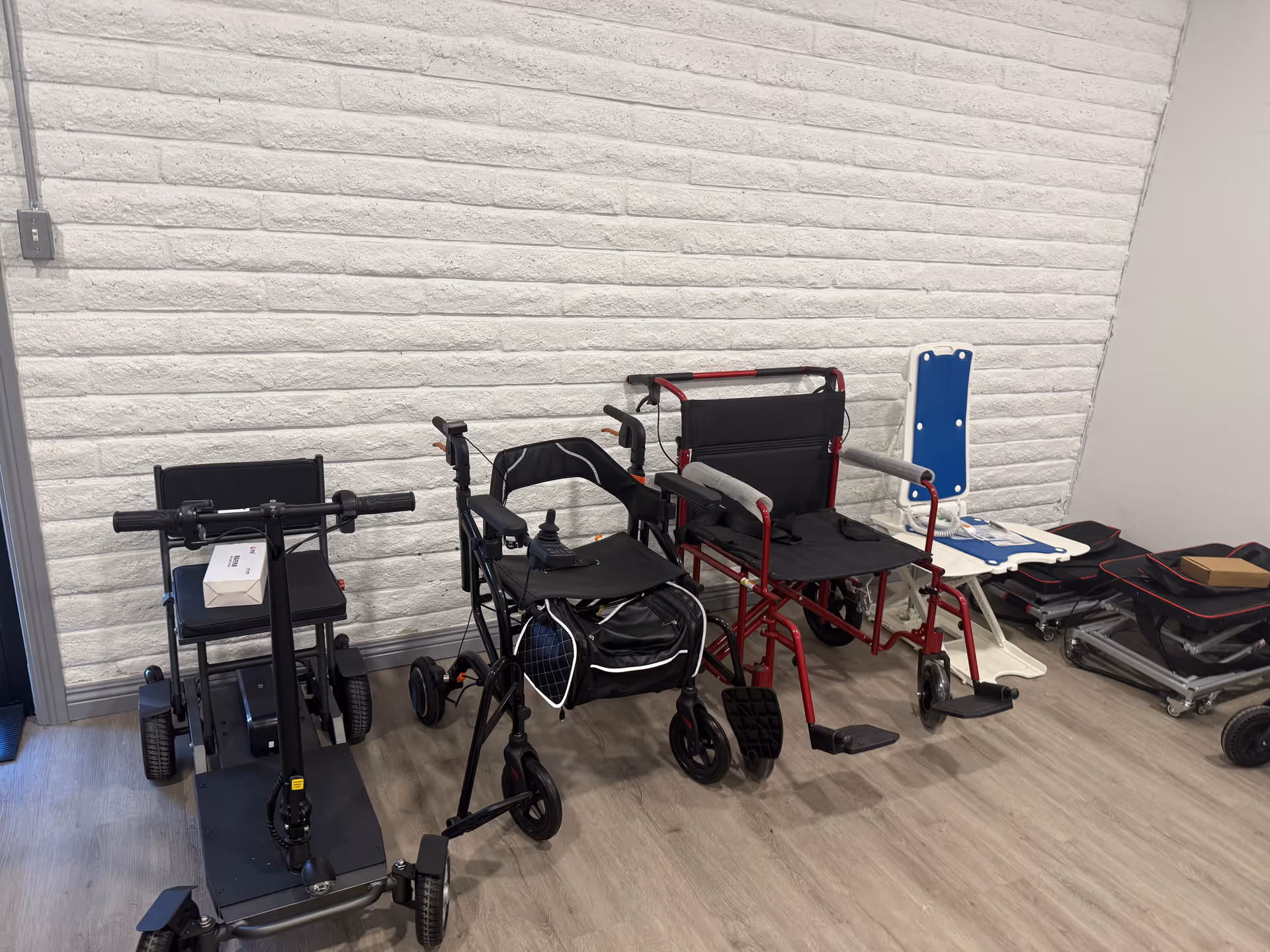 Several mobility aids like wheelchairs and walkers lined against a white brick wall indoors.