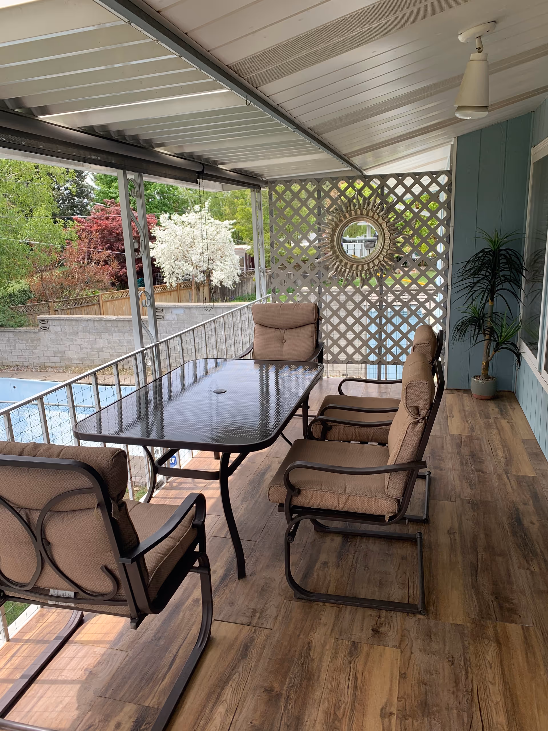 Covered outdoor patio with glass table, cushioned chairs, lattice wall, and a potted plant.