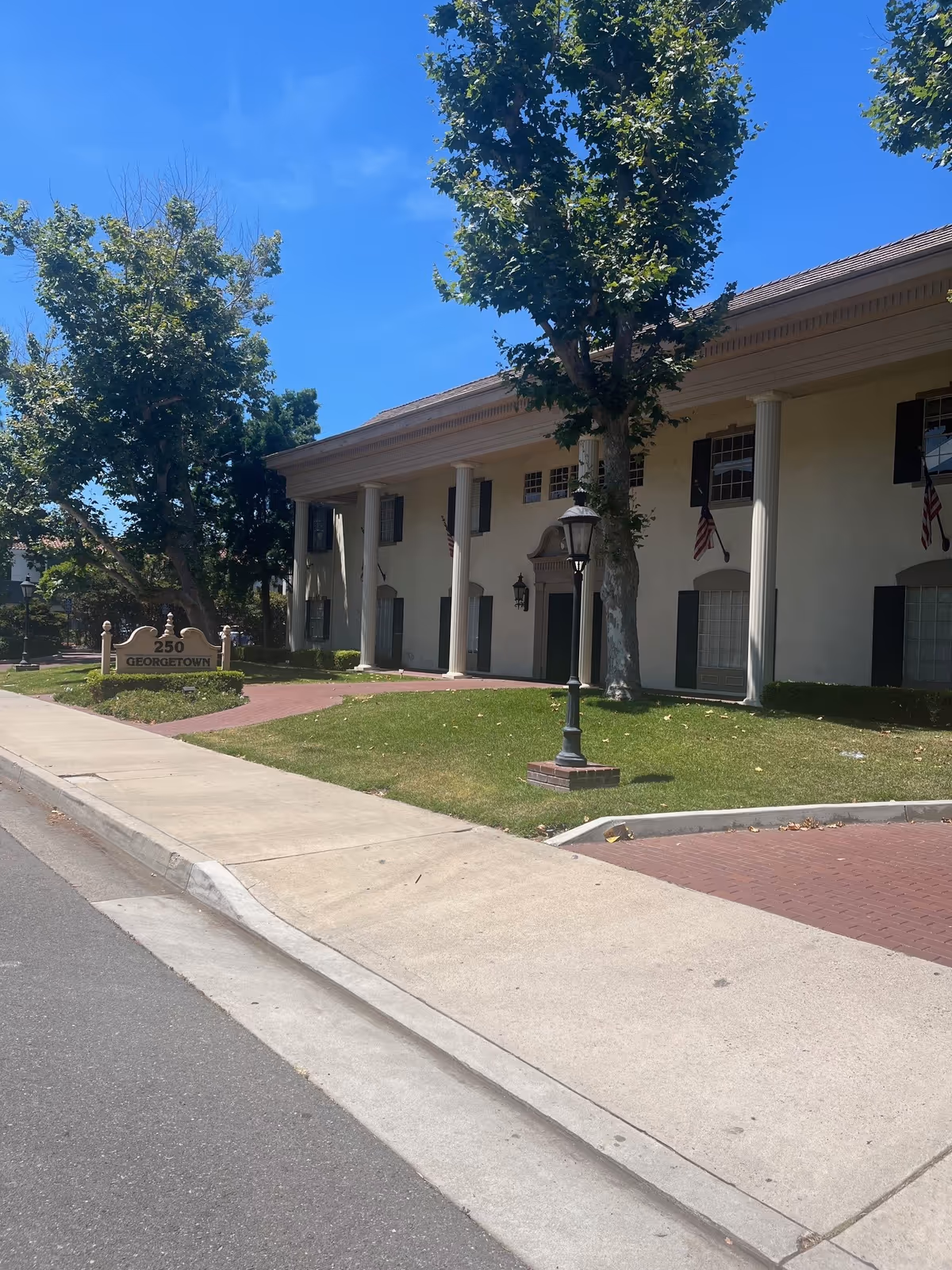 Front exterior of a senior living facility with columns, a sign, trees, and a sidewalk