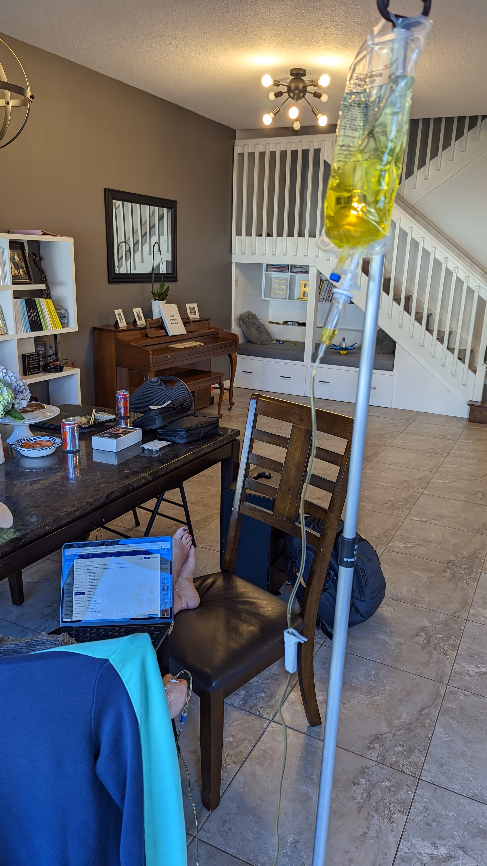Indoor living room with IV pole, dining table, laptop on resident, stairs in background.