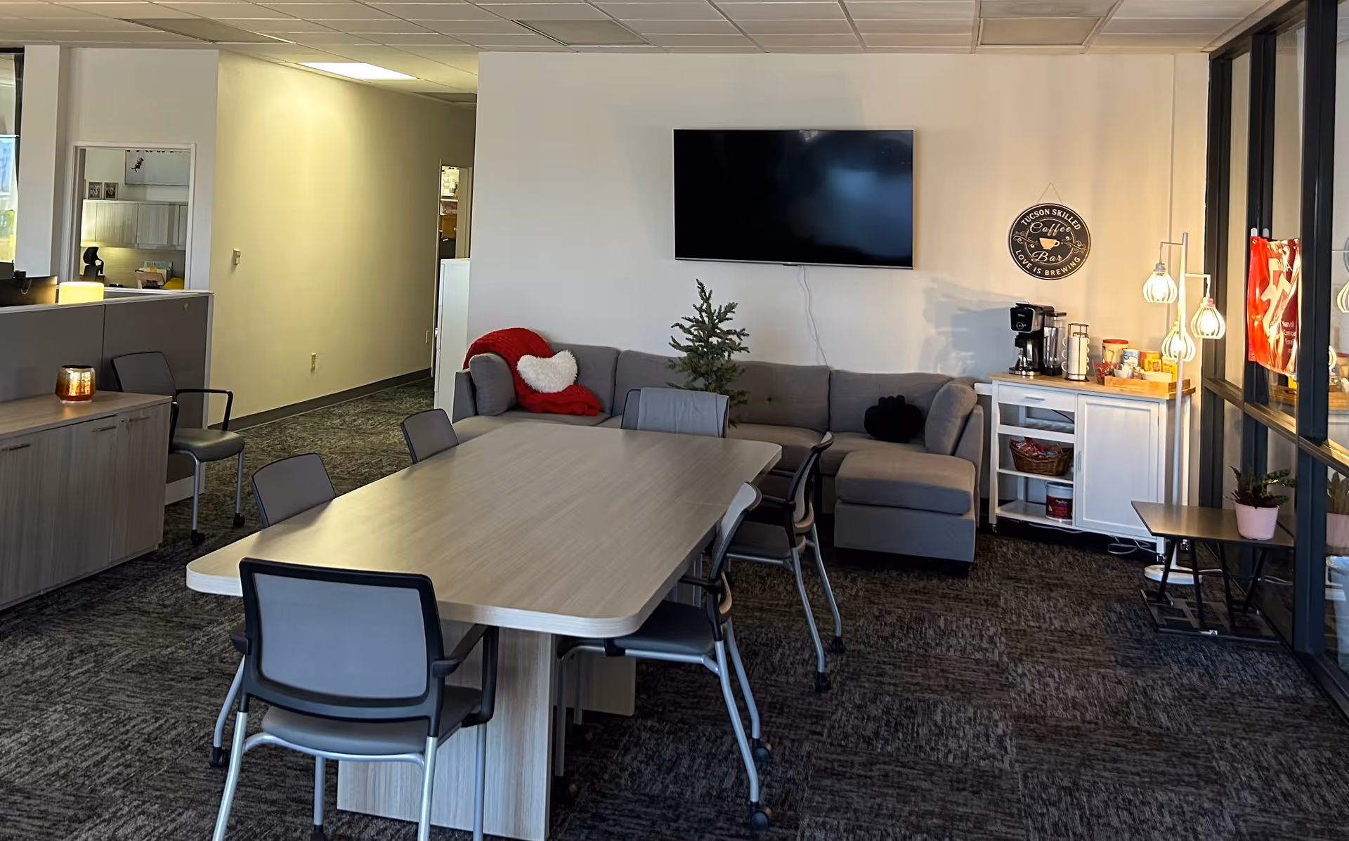 Bright shared living area with a large table, gray sofa, TV, and kitchenette.