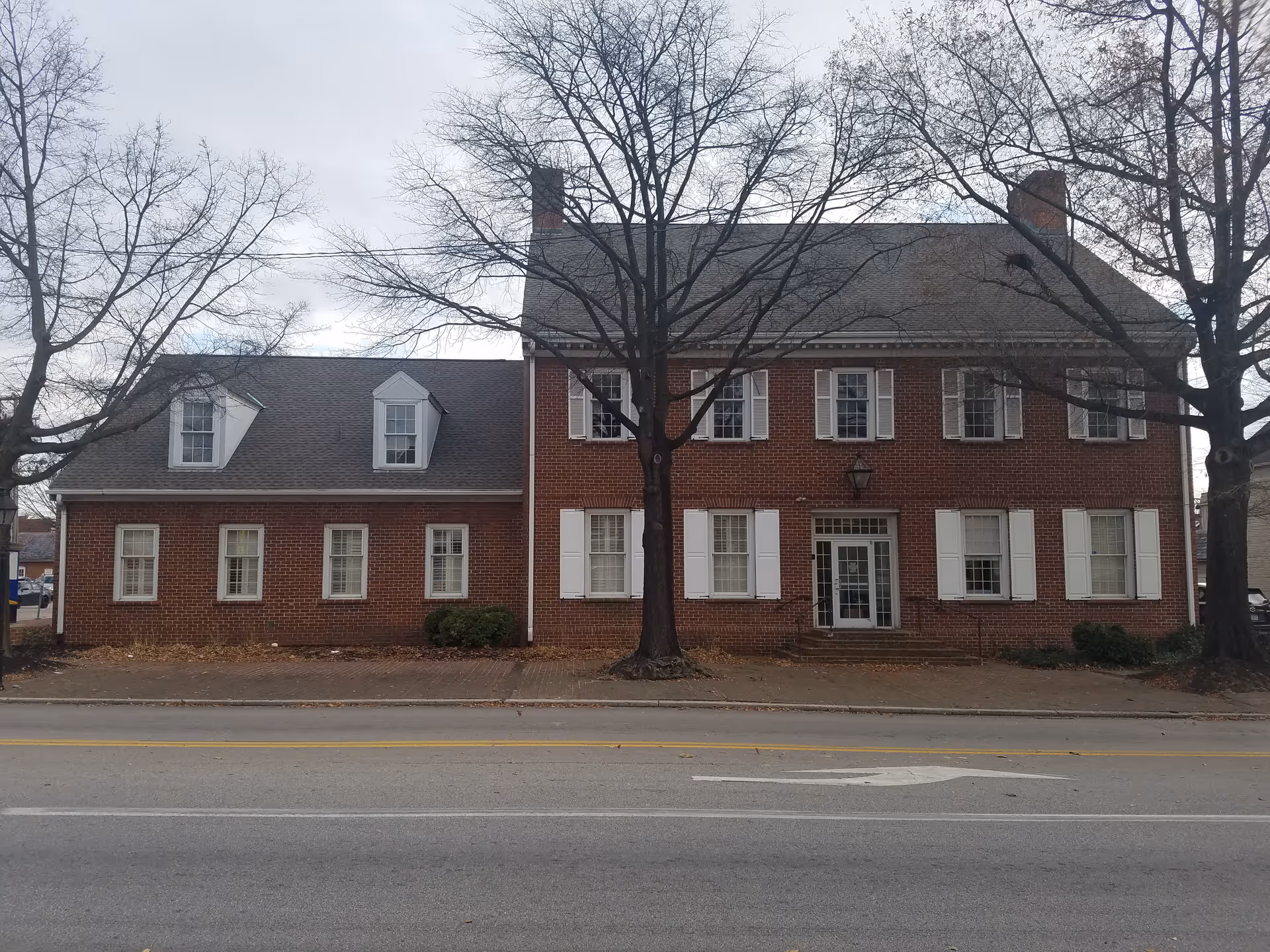 Brick, two-story building with white shutters along a street, leafless trees in front.