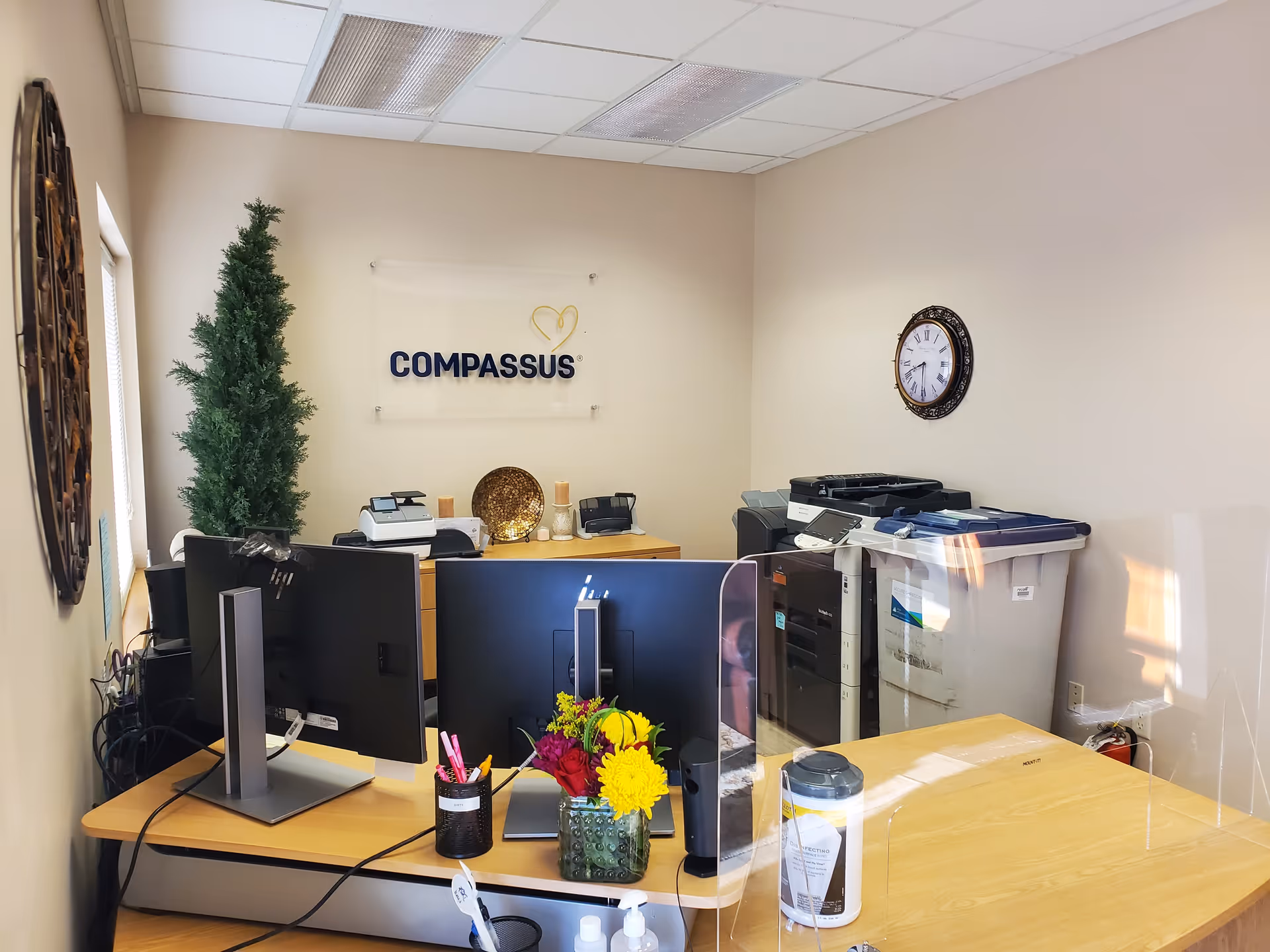 Office reception area with desks, computers, a Compassus sign, plant, and printer.
