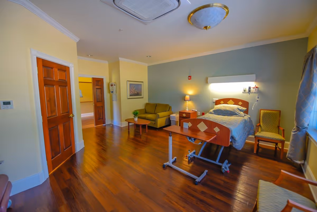 A bright hospice bedroom with a hospital bed, sofa, chairs, and wooden flooring.