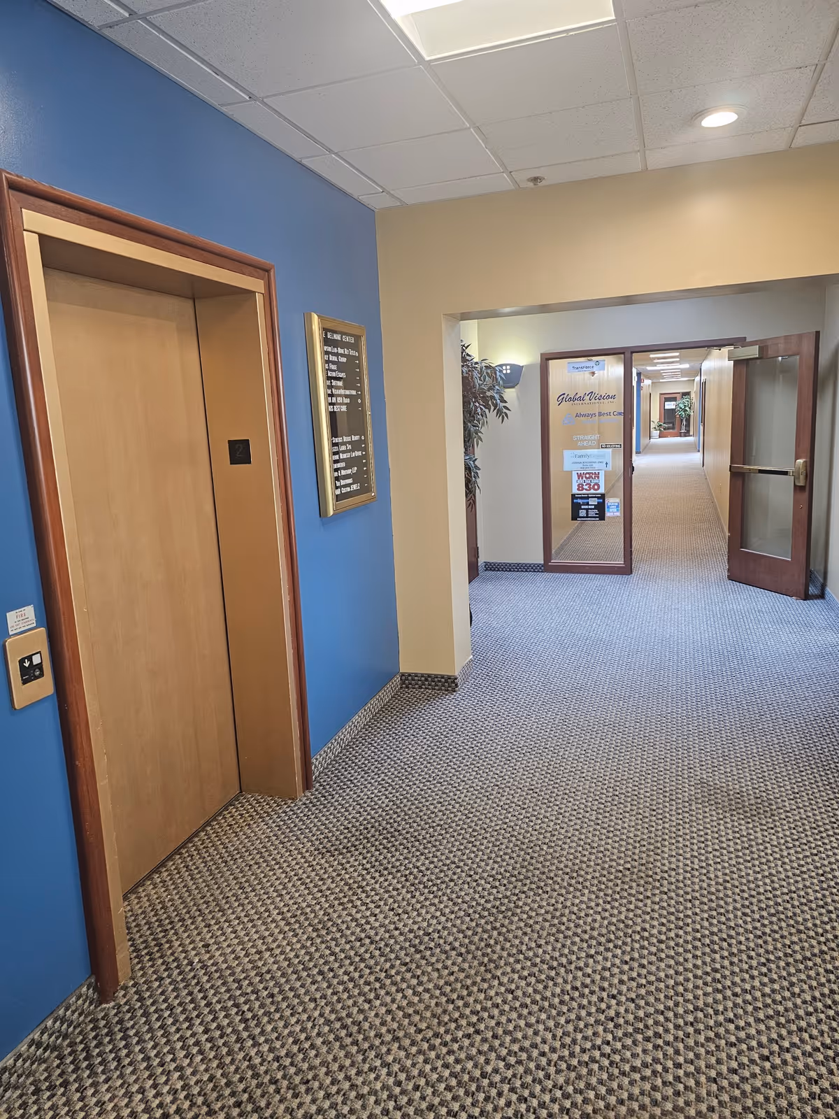 Indoor corridor with elevator on blue wall and open doors at hallway end.