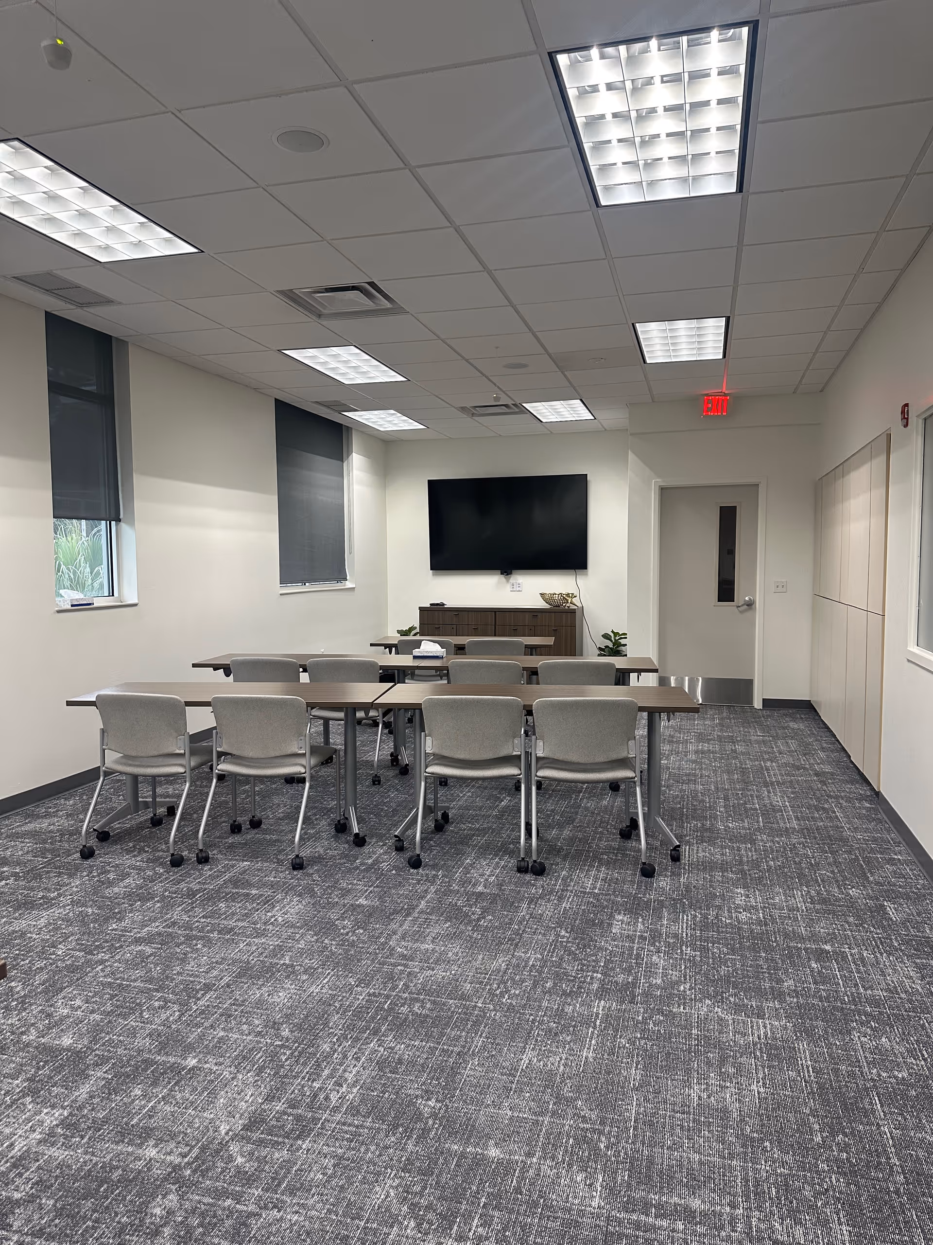 A conference room with tables, chairs, a wall-mounted TV, and windows.