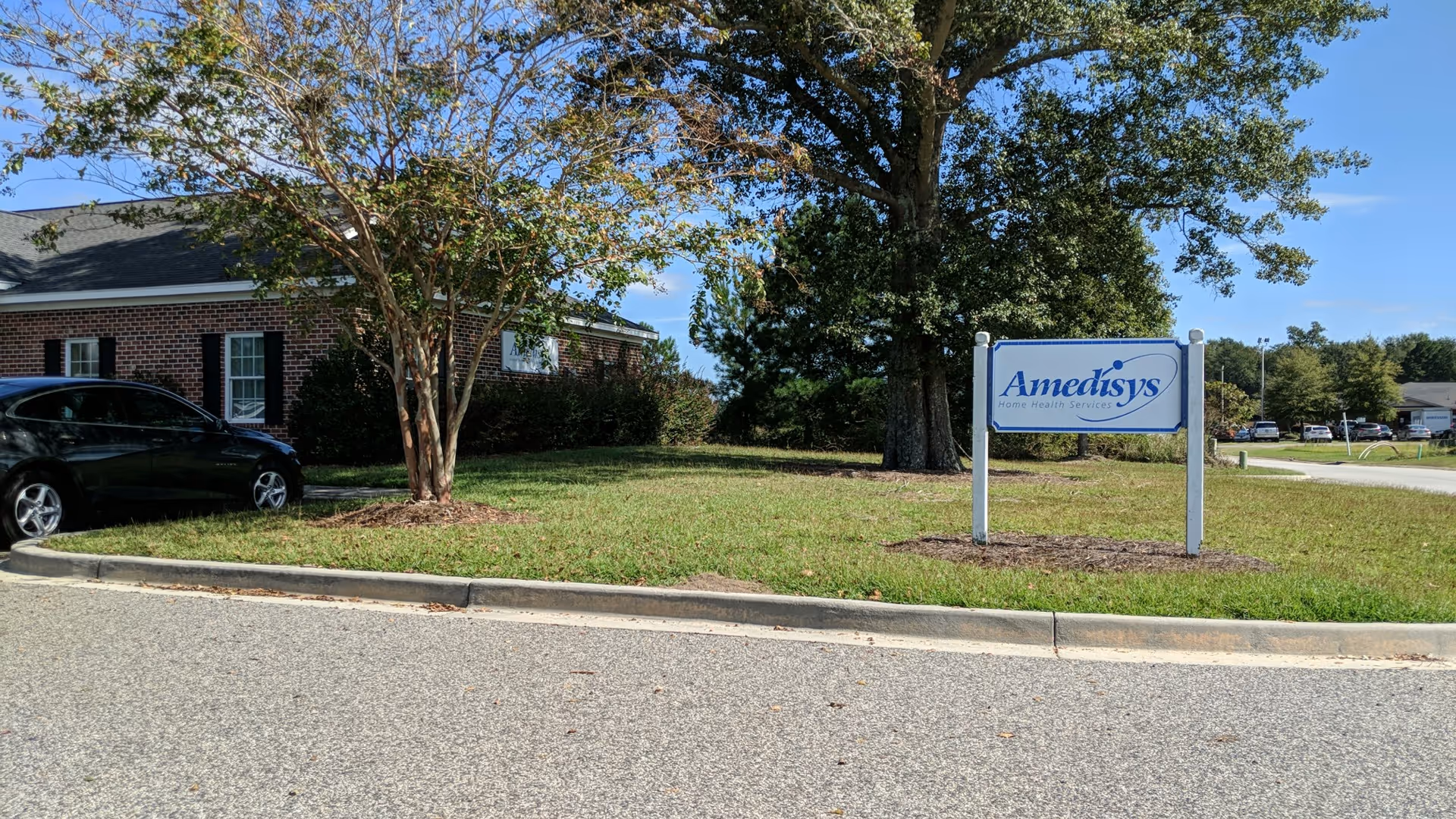Exterior view of a brick facility with an Amedisys sign, car, and green lawn.