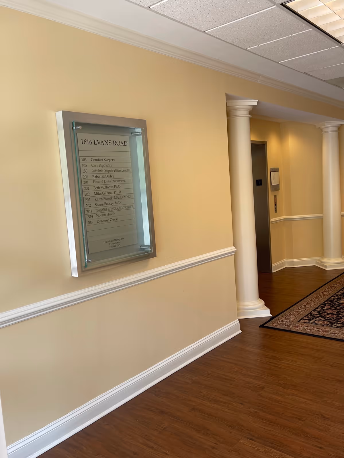 Interior hallway with a wall directory, beige walls, columns, and wood flooring.