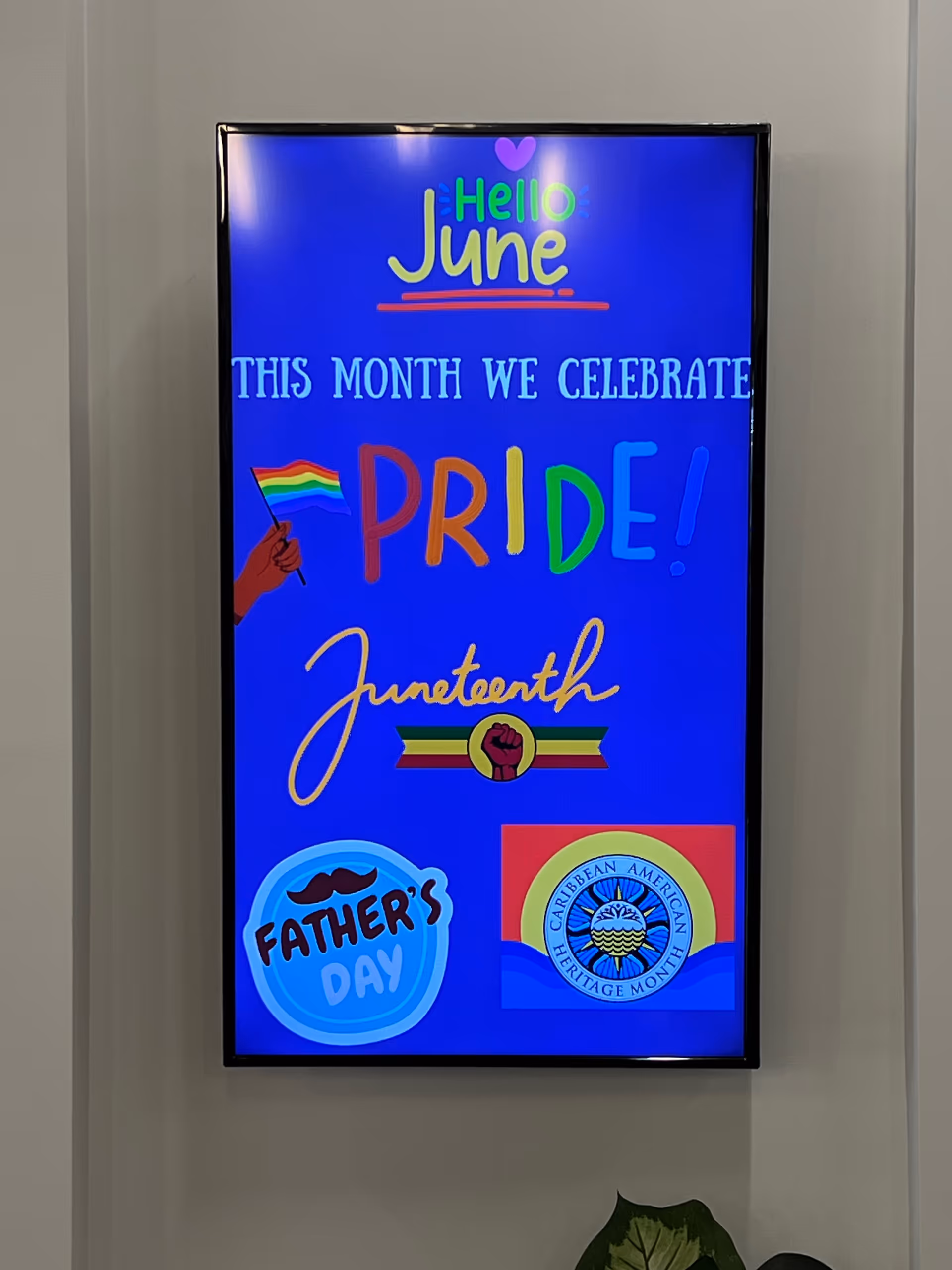 Bright vertical digital display in a hallway showing Pride month graphics and logos.