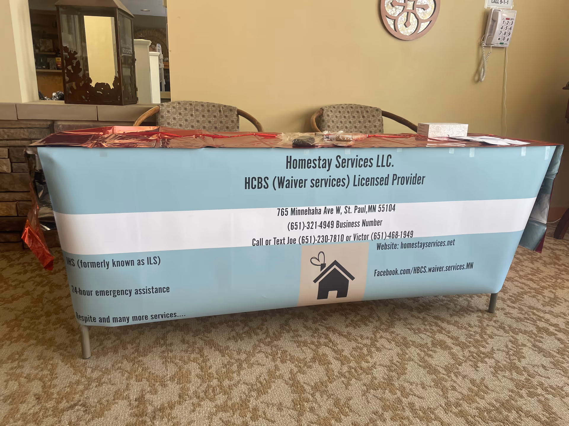 Banner-covered table in a lobby displays Homestay Services LLC with two chairs behind.