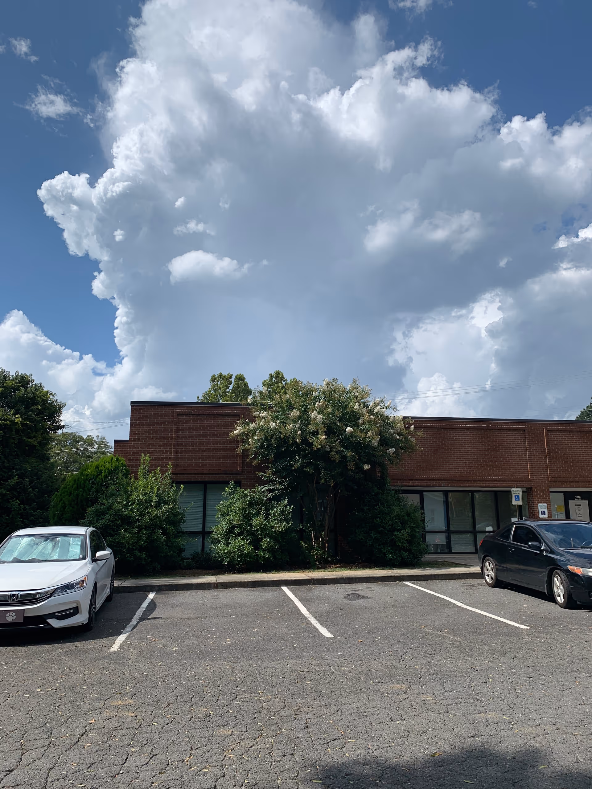 Brick facility with large windows, trees, and a parking lot in front of the building.