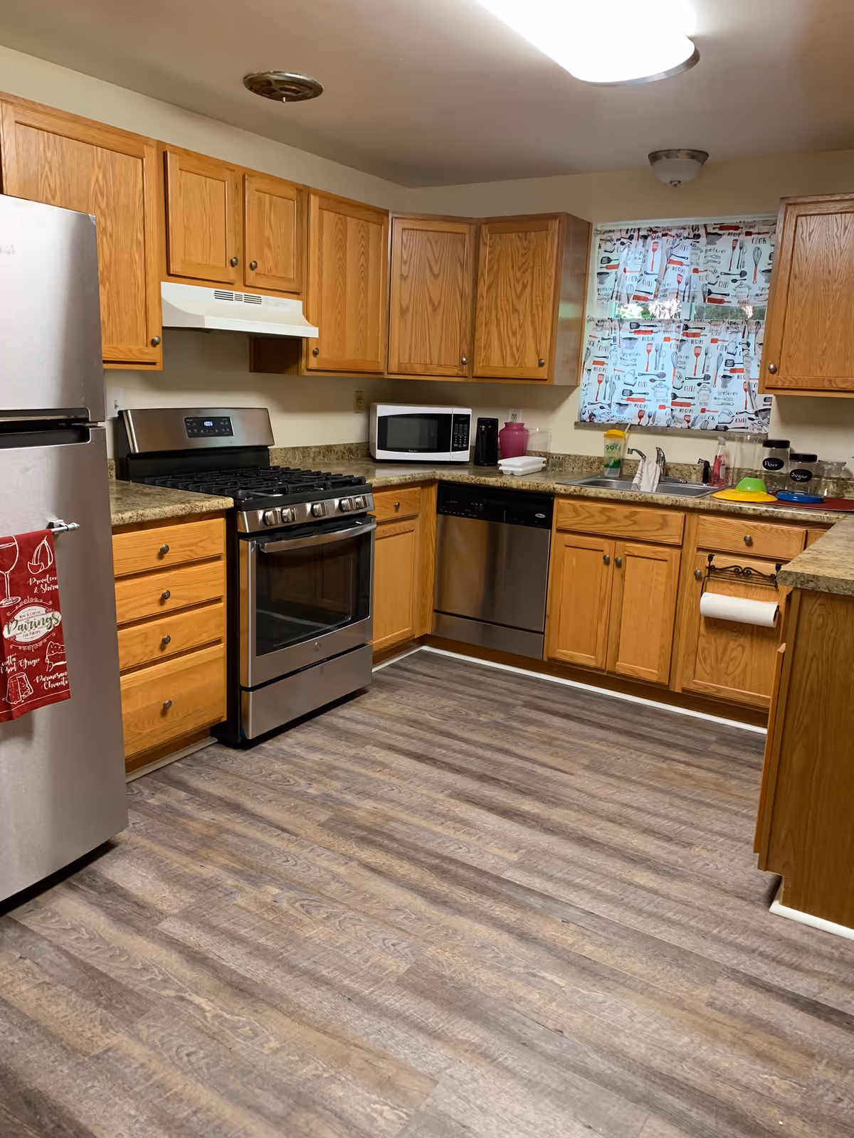Wooden kitchen cabinets surround stainless appliances and a double sink in an open facility kitchen.