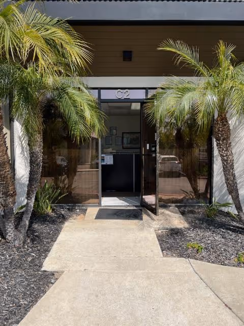Entrance with glass doors opening to a lobby, framed by palm trees and a concrete path.