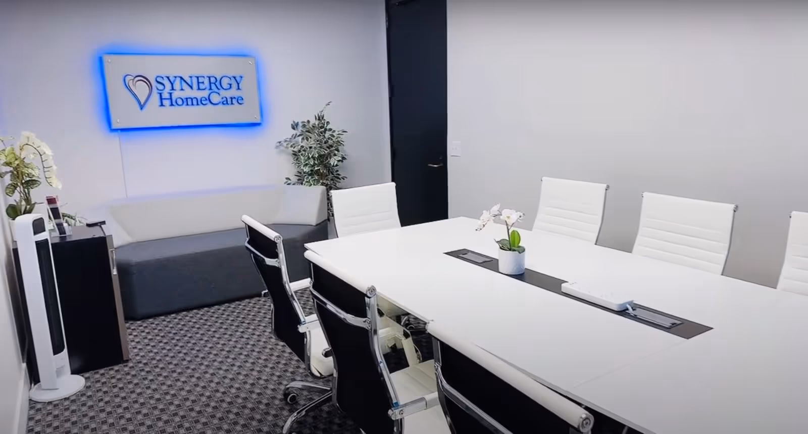 Modern conference room with white table, white chairs, plants, and a Synergy HomeCare sign.