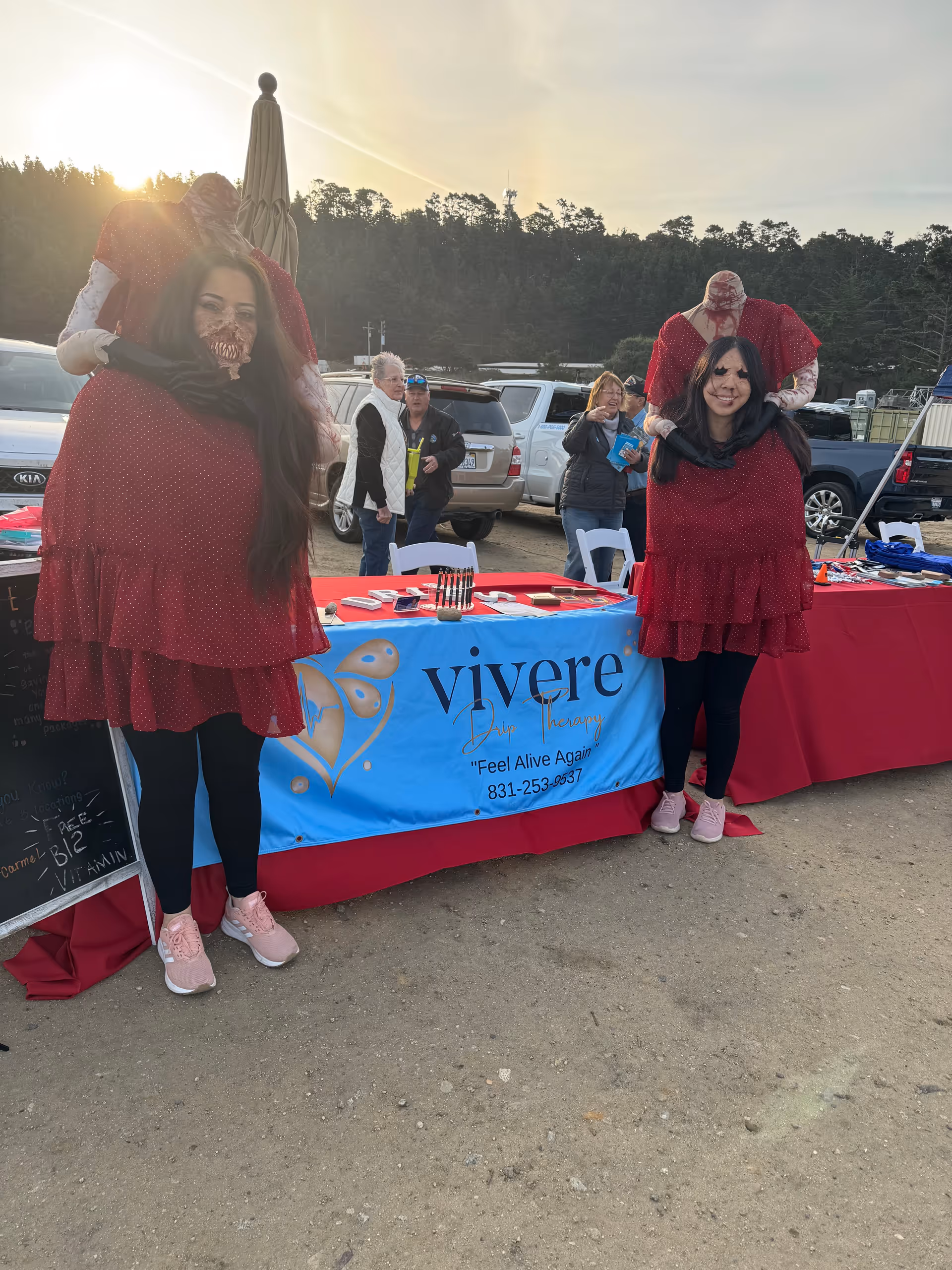Two women in red dresses stand by a Vivere Drip Therapy banner at an outdoor event.