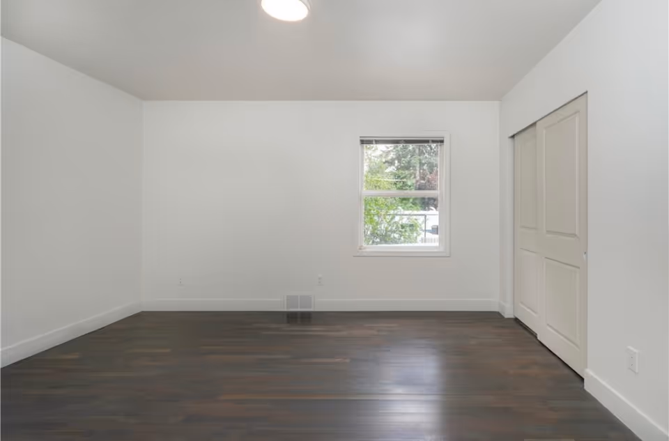 A small empty white-painted room with a window, closet doors, and dark wood floors.