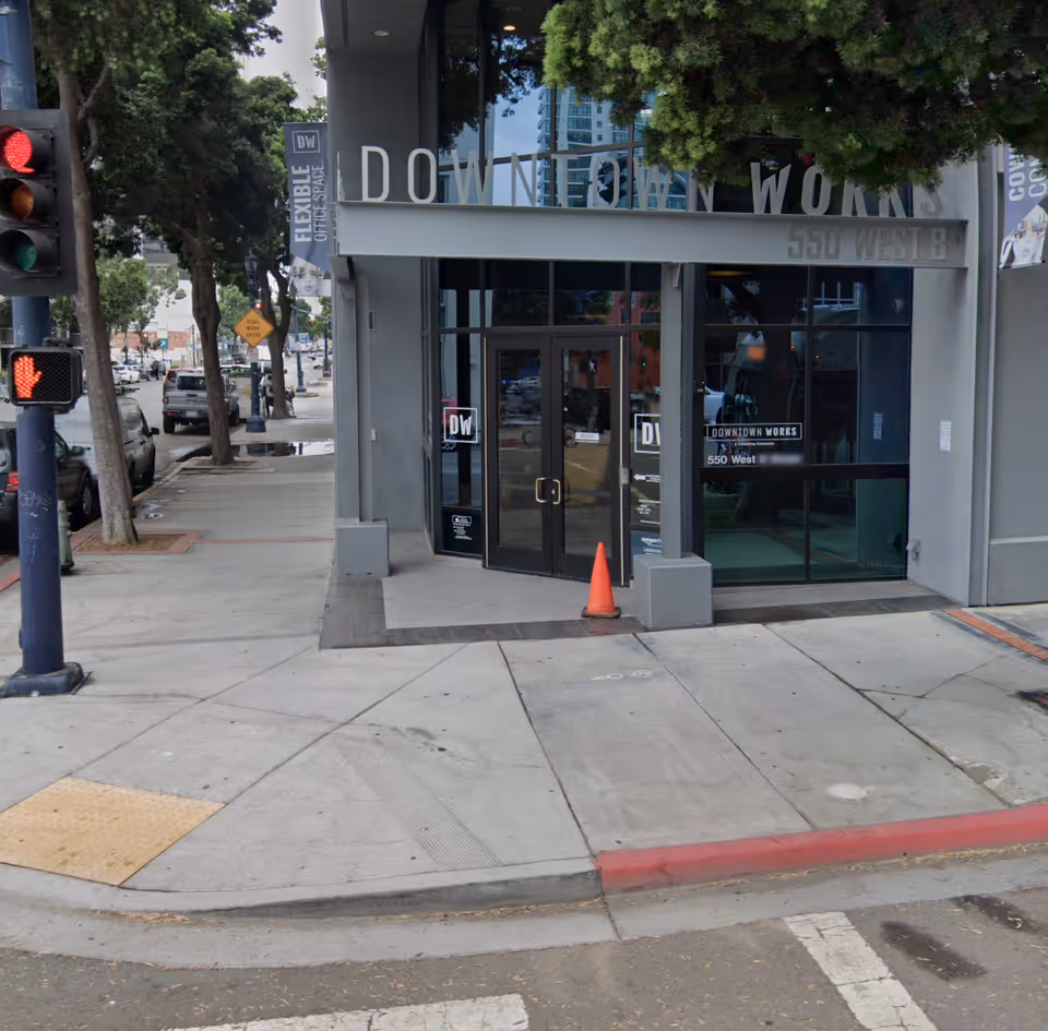 City sidewalk with a modern storefront entrance labeled Downtown Works, glass doors, trees, and street scene.