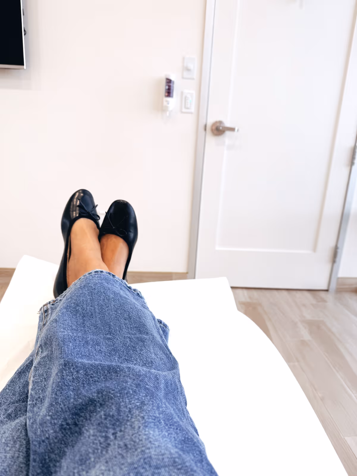 View from a patient bed showing legs in jeans inside a clinic room.