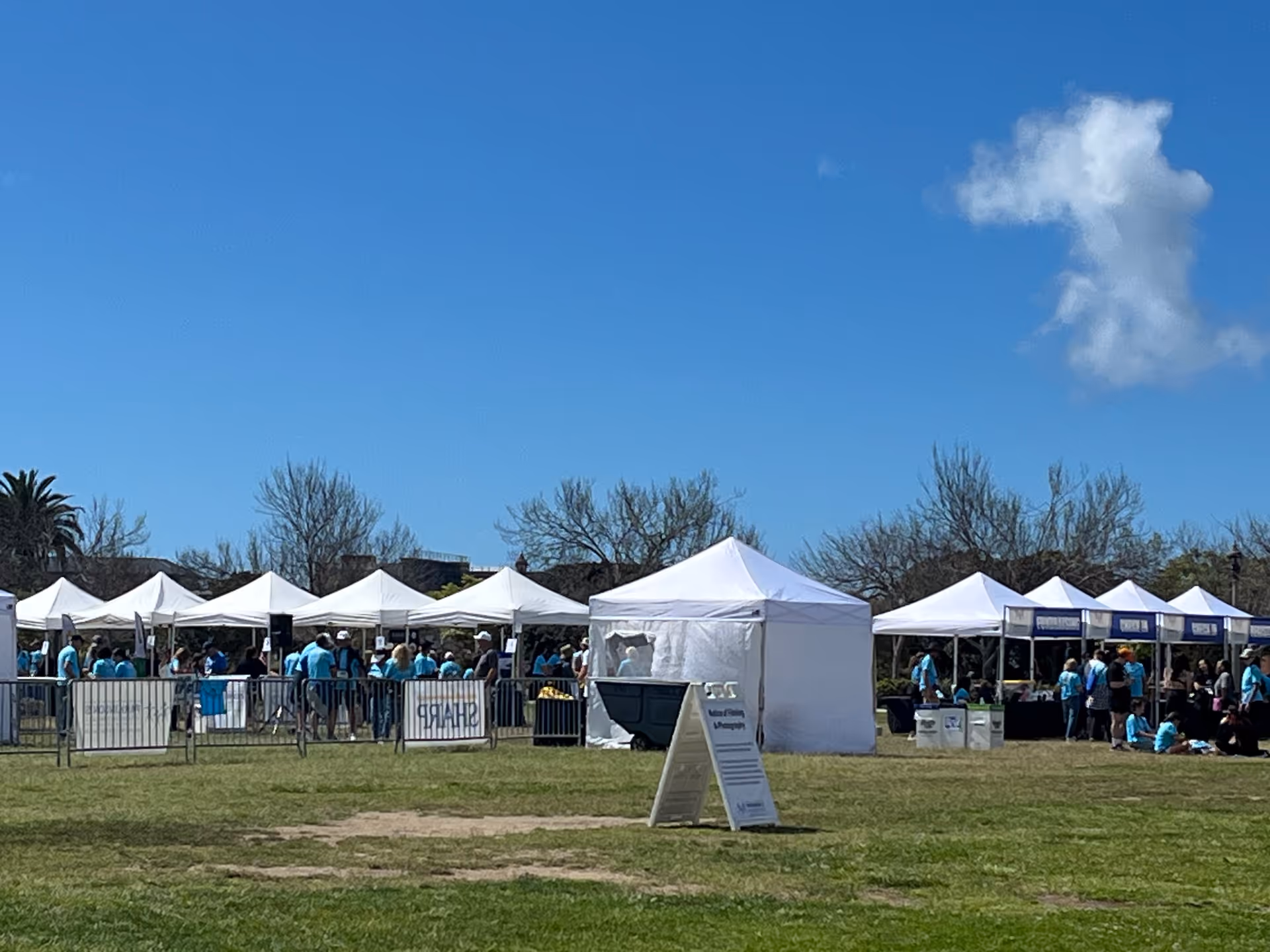 Row of white tents and people outdoors on a sunny day at a community event.