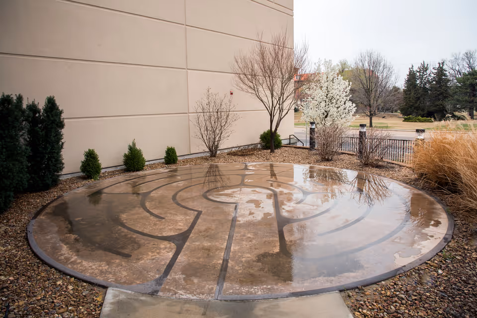 A circular metal maze design on a wet outdoor patio beside a beige building.