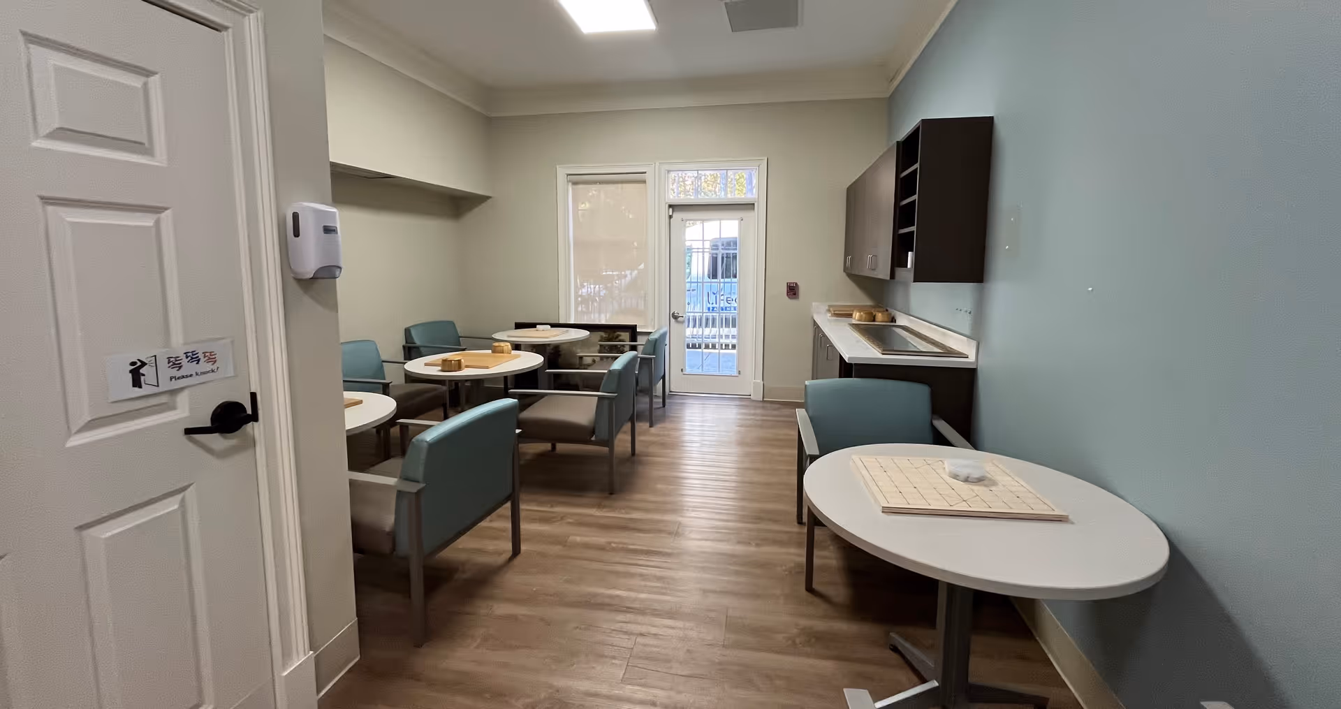 Senior living dining area with round tables, teal chairs, and kitchenette.