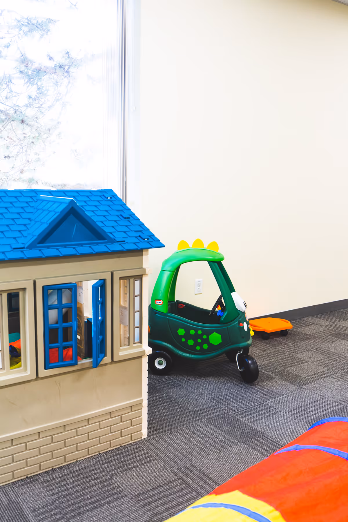 Indoor play area with a plastic toy house, green ride-on car, and colorful mat.