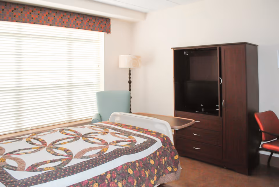 Bedroom in a senior living facility with a quilted bed, chair, and TV cabinet.