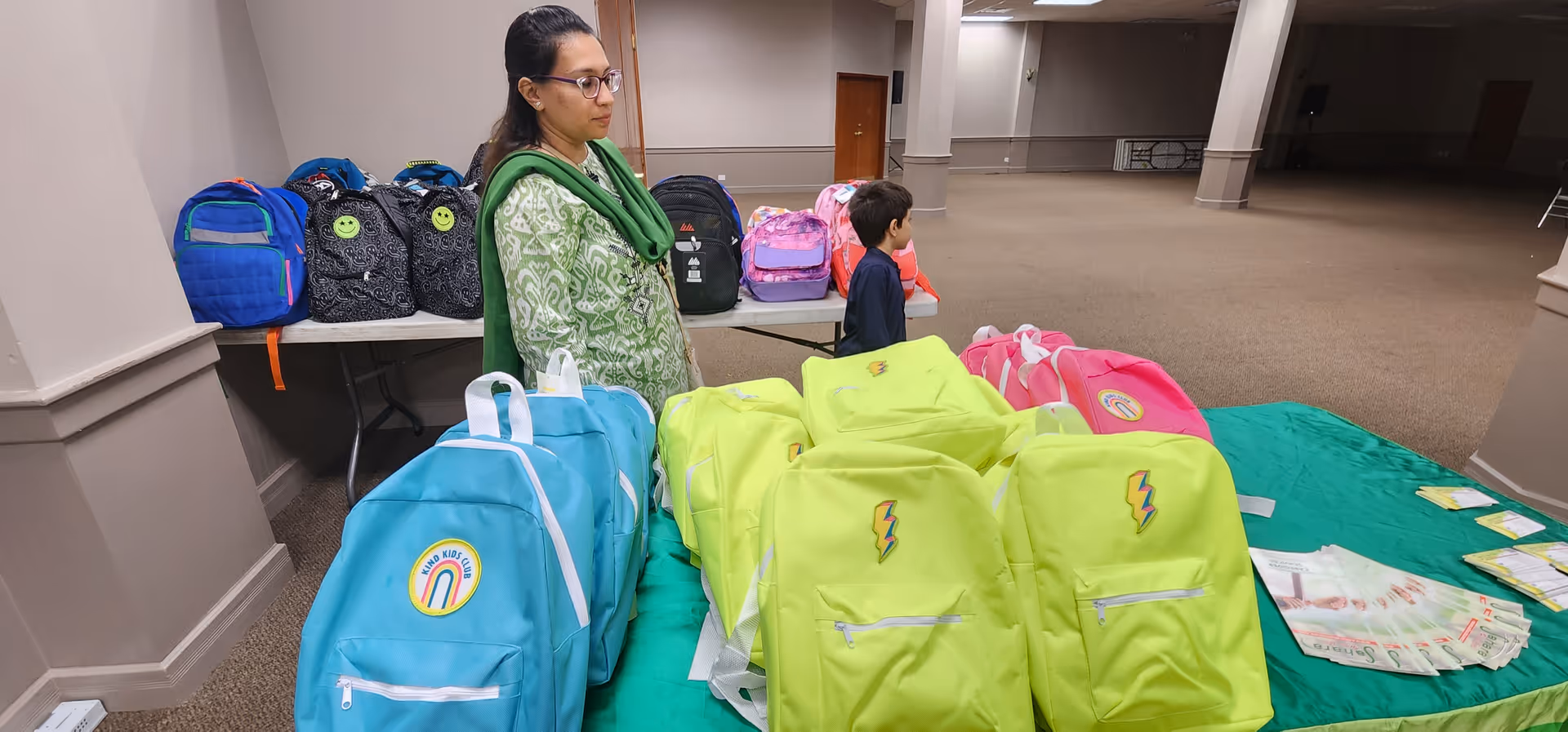 A woman and a child stand near a table of colorful backpacks in spacious indoor hallway.