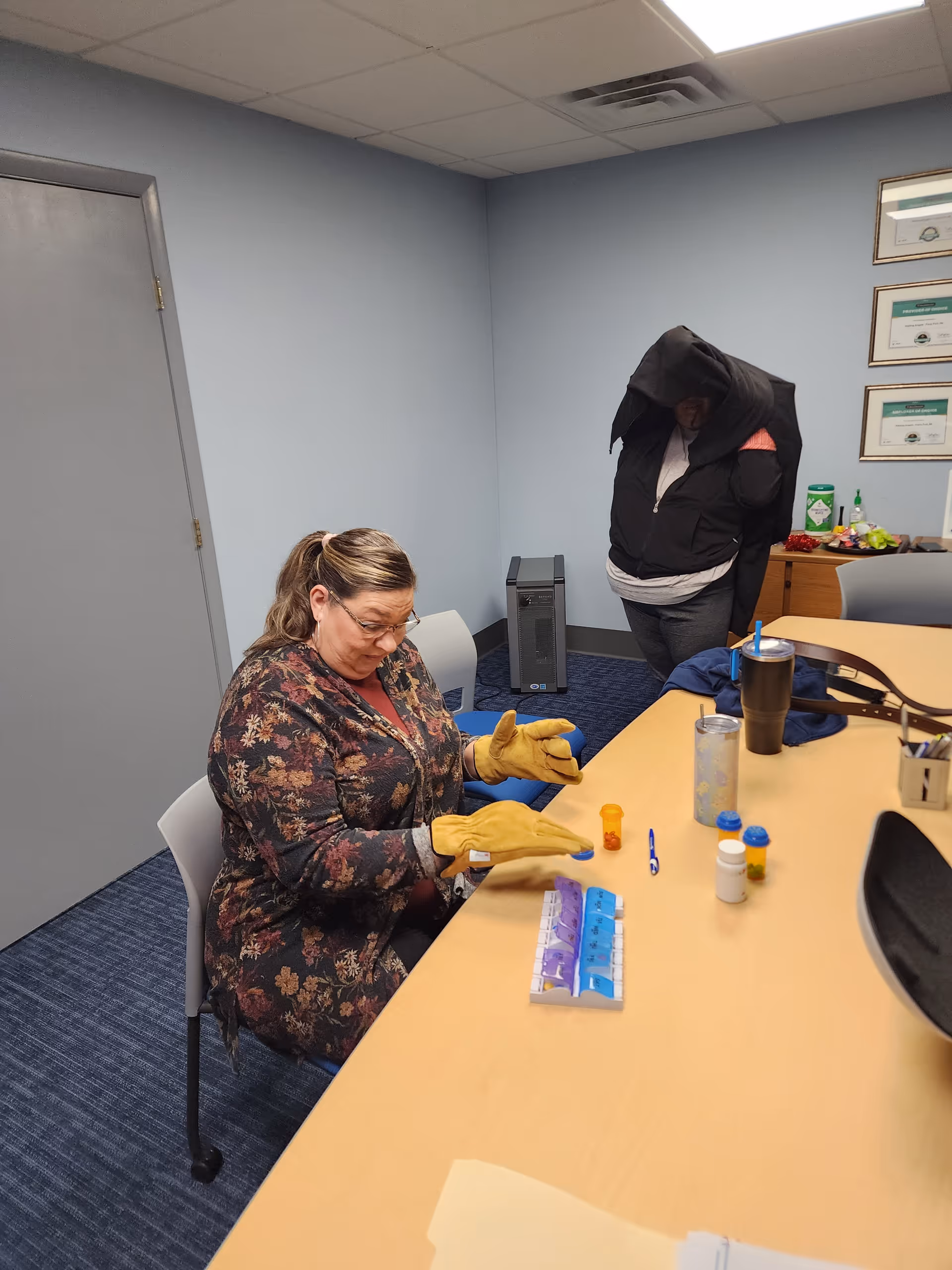 Caregiver wearing gloves sits at a table with medication supplies, while another person stands nearby.