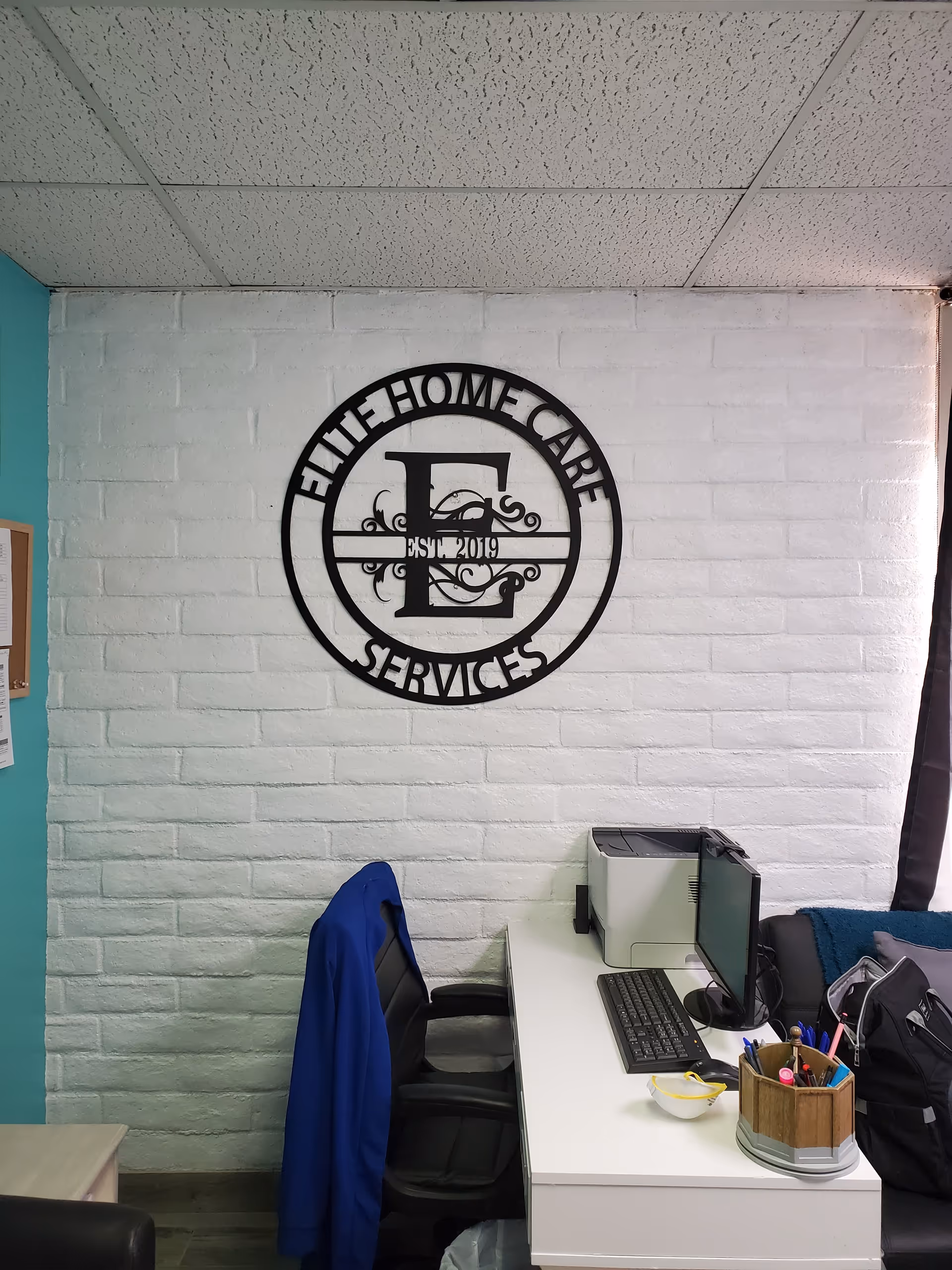Office desk with computer and jacket in front of Elite Home Care Services logo on wall.