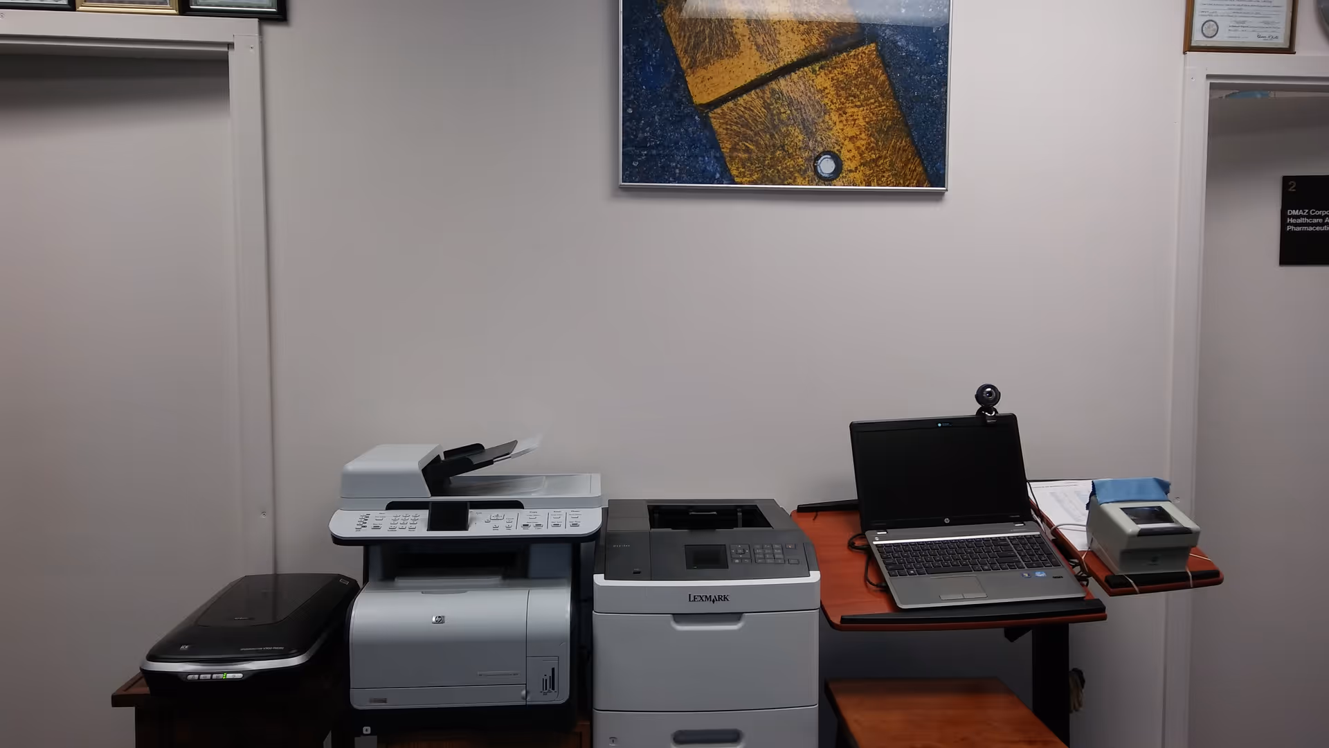 Interior office area with a printer, copier, laptop, webcam, and office supplies on a red table.