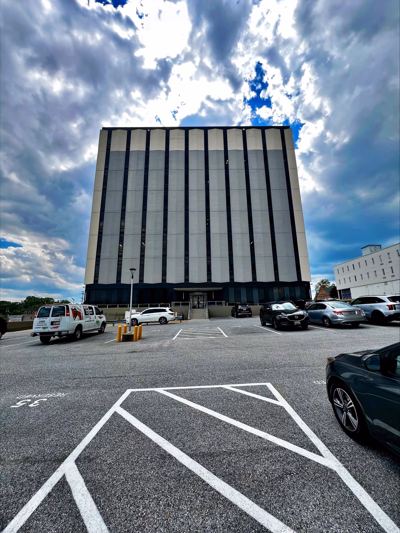 Exterior of a tall modern building with a parking lot and cloudy sky.