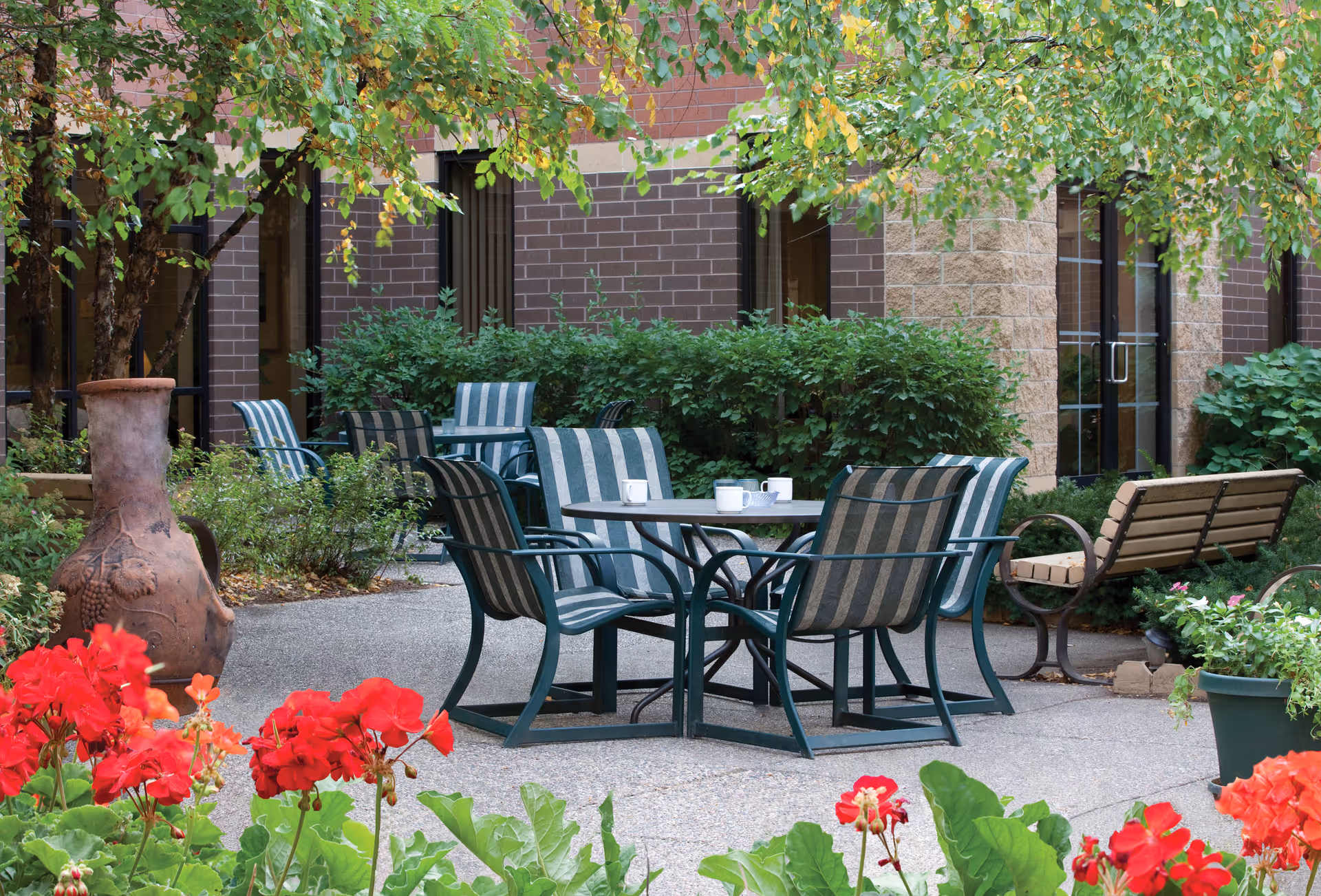 Outdoor courtyard with metal tables, striped chairs, benches, potted plants, and brick building.