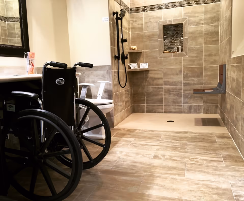 Accessible bathroom with wheelchair, tiled shower, grab bars, and toilet in a modern facility.