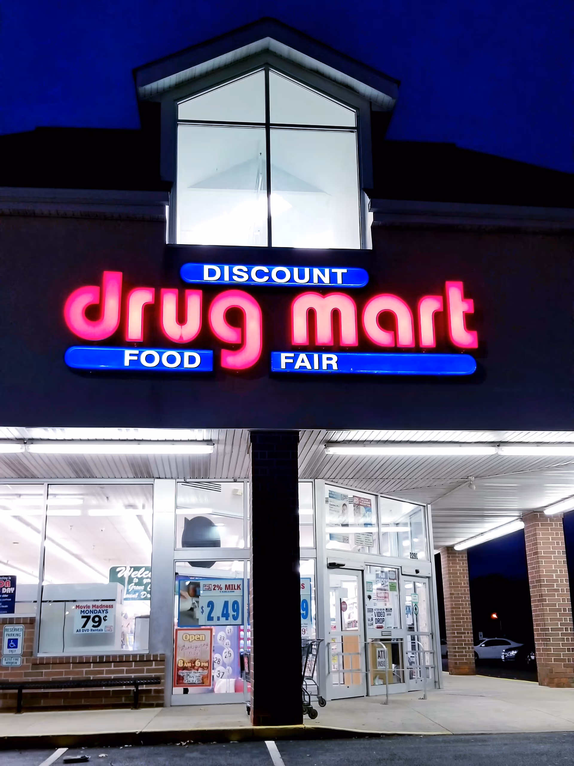 Nighttime storefront with neon Discount Drug Mart sign and glass entry doors.