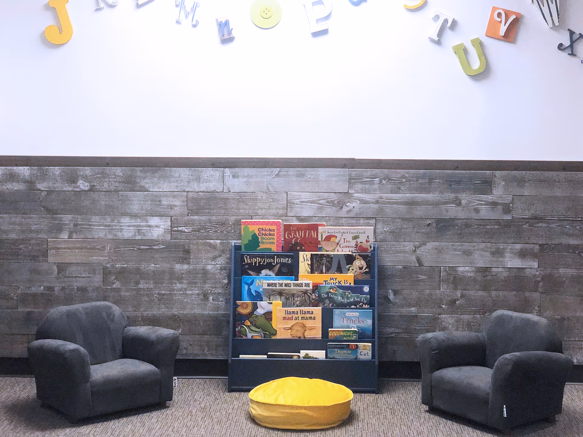 Two gray armchairs flank a bookshelf with children's books in a cozy lobby.